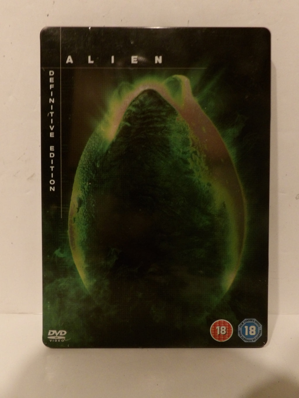 Alien Definitive Edition DVD steelbook case featuring the iconic green bioweapon graphic on a black background, with 18 rating logos in the bottom right corner.