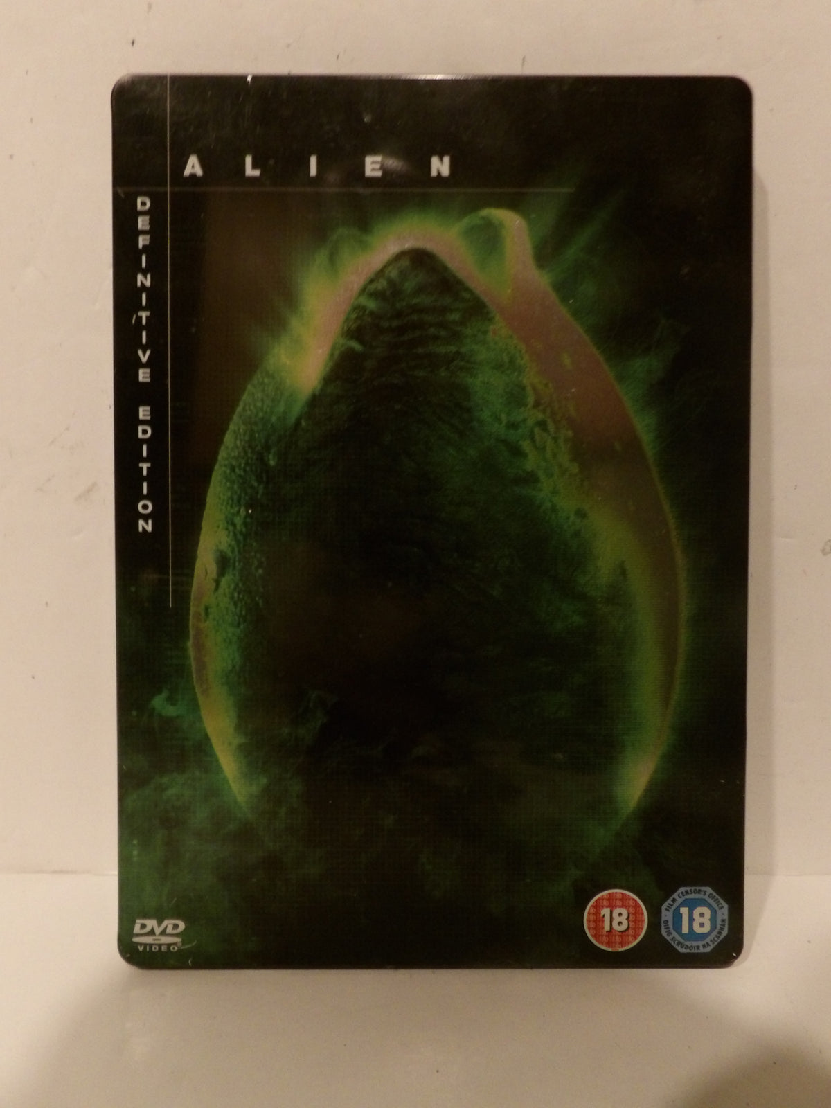 Alien Definitive Edition DVD steelbook case featuring the iconic green bioweapon graphic on a black background, with 18 rating logos in the bottom right corner.