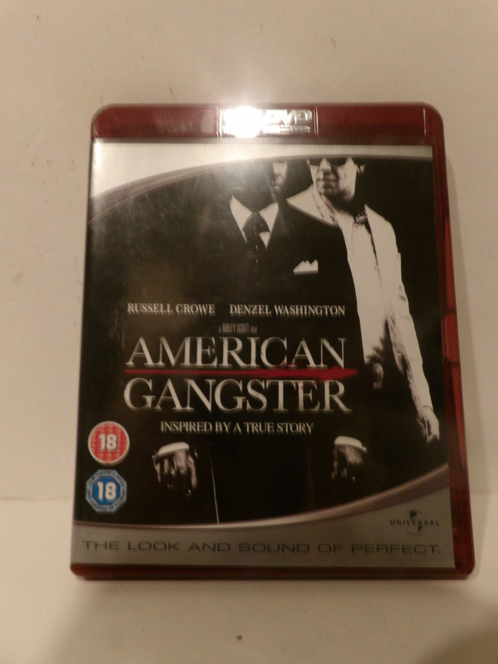 A boxed movie case for 'American Gangster' with Russell Crowe and Denzel Washington, photographed against a plain off-white background. The cover is mostly black and white with red accents, and age rating badges are visible on the lower left.