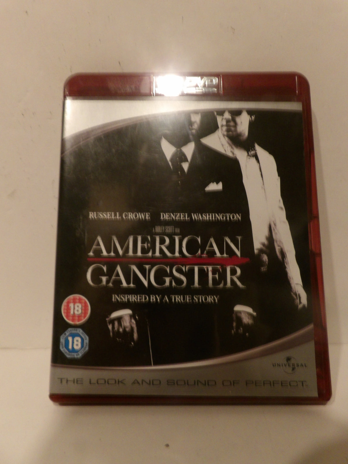 A boxed movie case for 'American Gangster' with Russell Crowe and Denzel Washington, photographed against a plain off-white background. The cover is mostly black and white with red accents, and age rating badges are visible on the lower left.