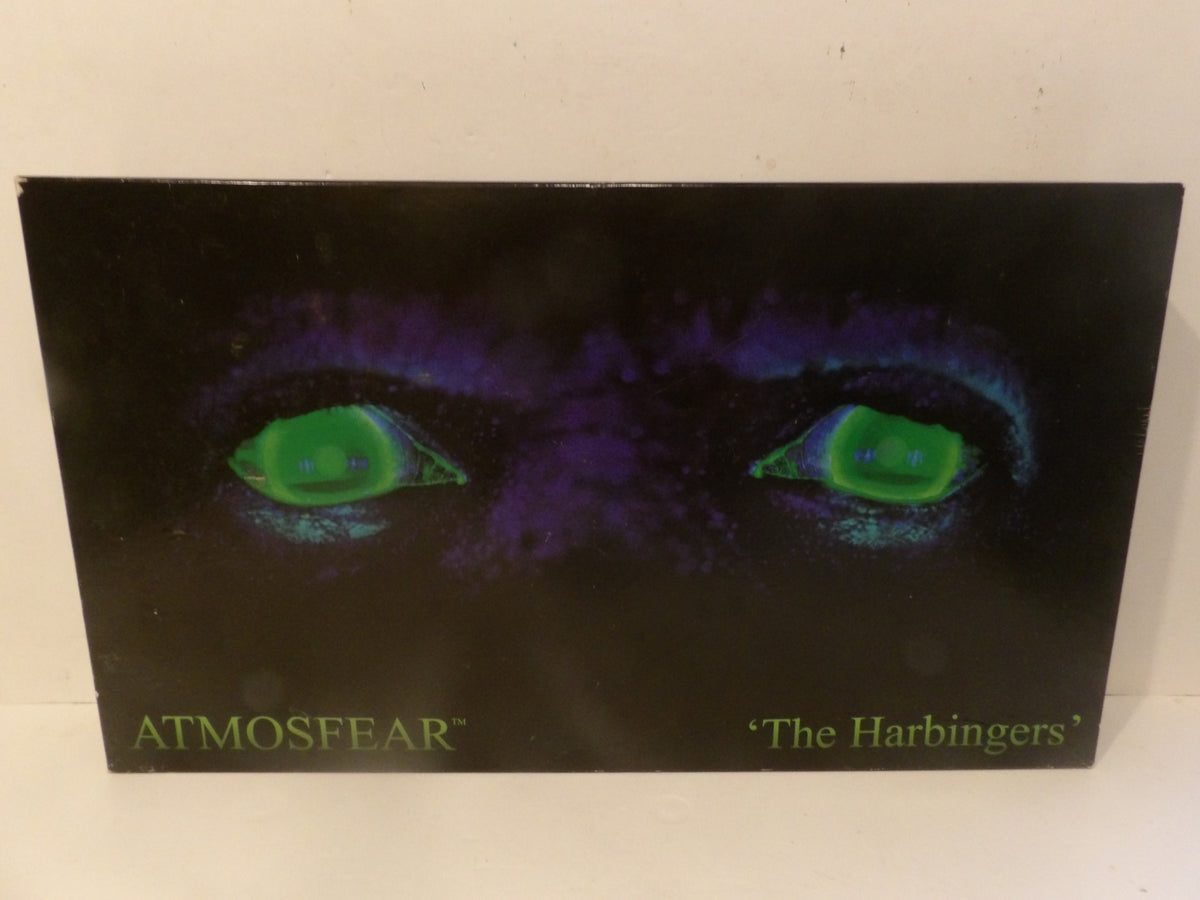 A square album cover for Atmosphere's 'The Harbingers' featuring a dark, eerie design with glowing green eyes on a black and purple background, photographed against a light off-white surface.
