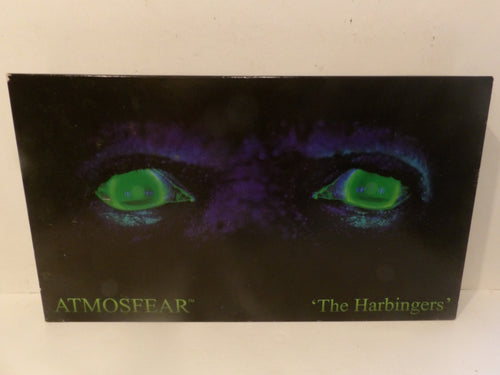A square album cover for Atmosphere's 'The Harbingers' featuring a dark, eerie design with glowing green eyes on a black and purple background, photographed against a light off-white surface.