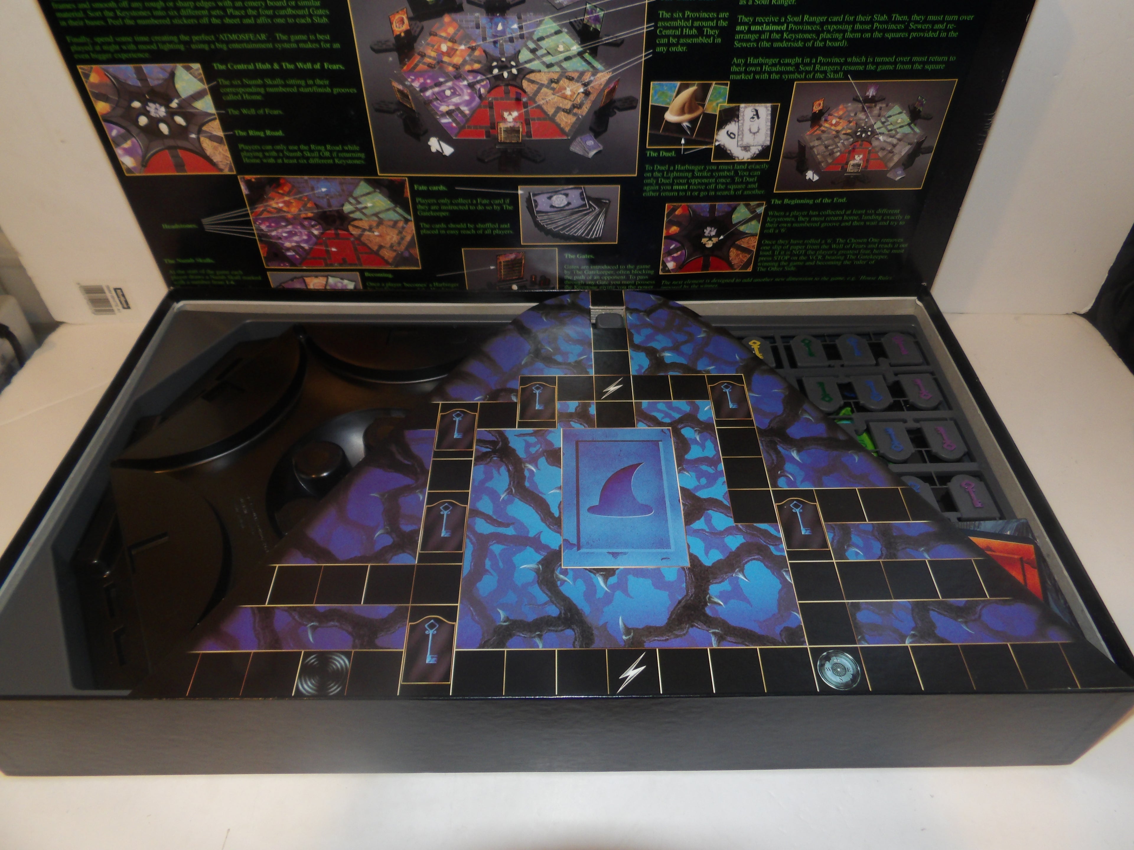 A board game set displayed open in its box, featuring a dark fantasy-themed board with blue and purple artwork, black playmat sections, and various game pieces. The inside lid contains printed rules or artwork panels.