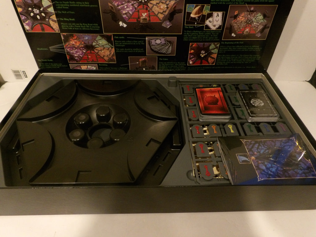 Open board game box showing the game components, including a large black circular piece, cards, tokens, and a small illustrated booklet on a white surface.