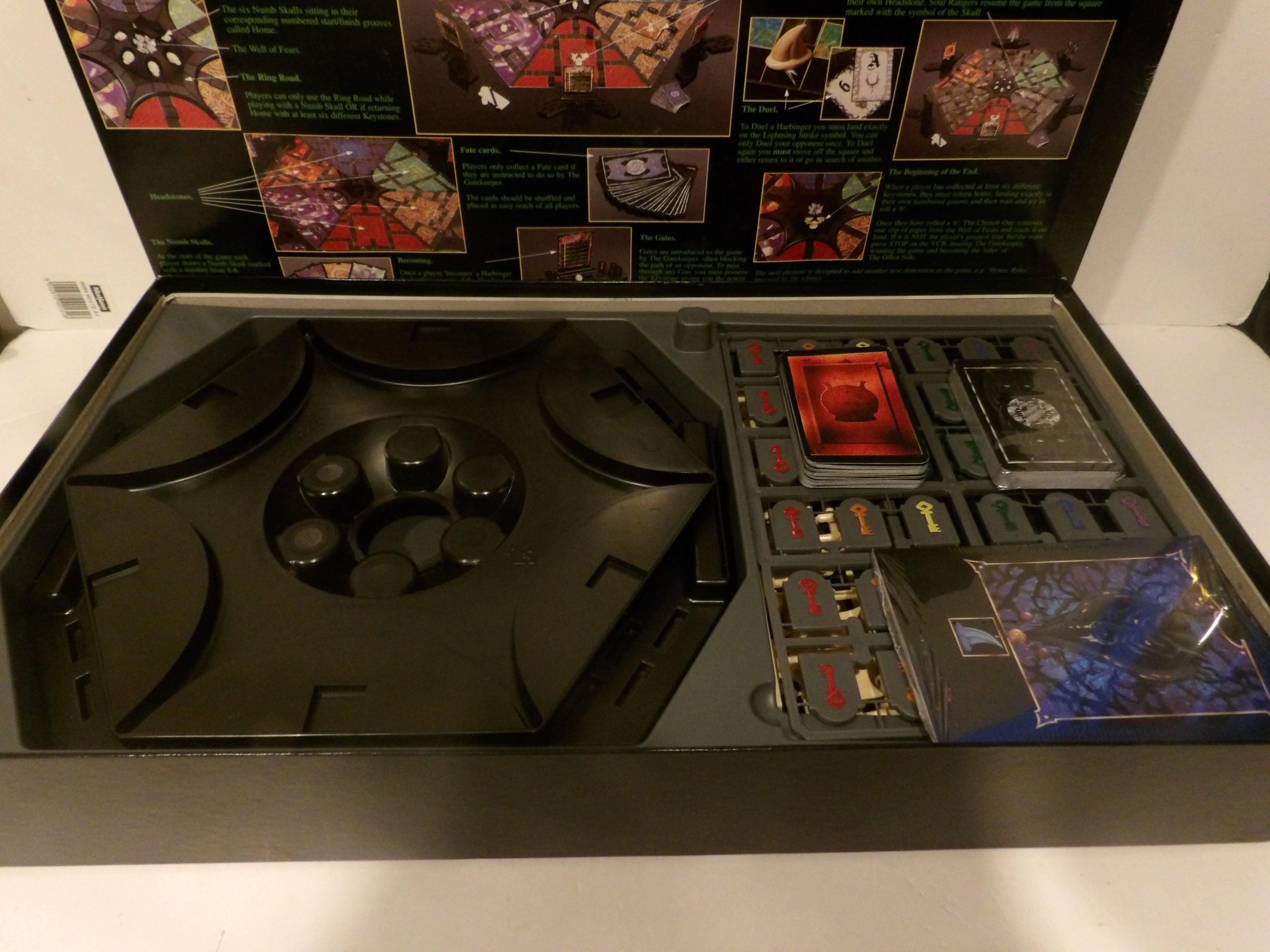 Open board game box showing the game components, including a large black circular piece, cards, tokens, and a small illustrated booklet on a white surface.