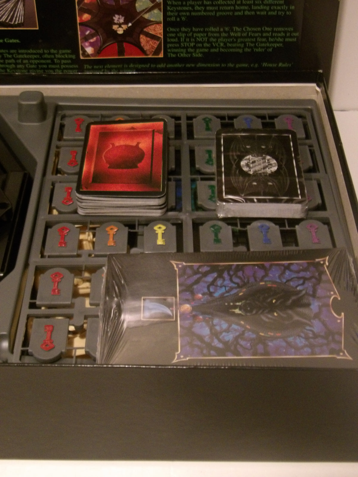 A boxed board game set opened to reveal a gray game tray with multiple cards, tokens, and a clear plastic-wrapped component. The packaging insert at the top shows illustrated artwork and text, but the main visible subject is the opened game box and its contents.