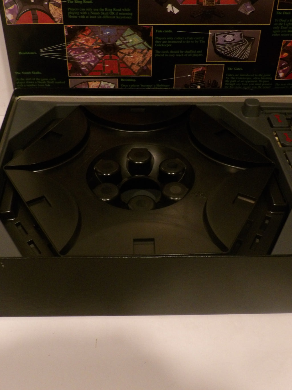 A black plastic game board tray or playmat shown open, with printed game artwork visible along the inner back panel. The tray has molded circular and angular sections and appears to be part of a tabletop game set.