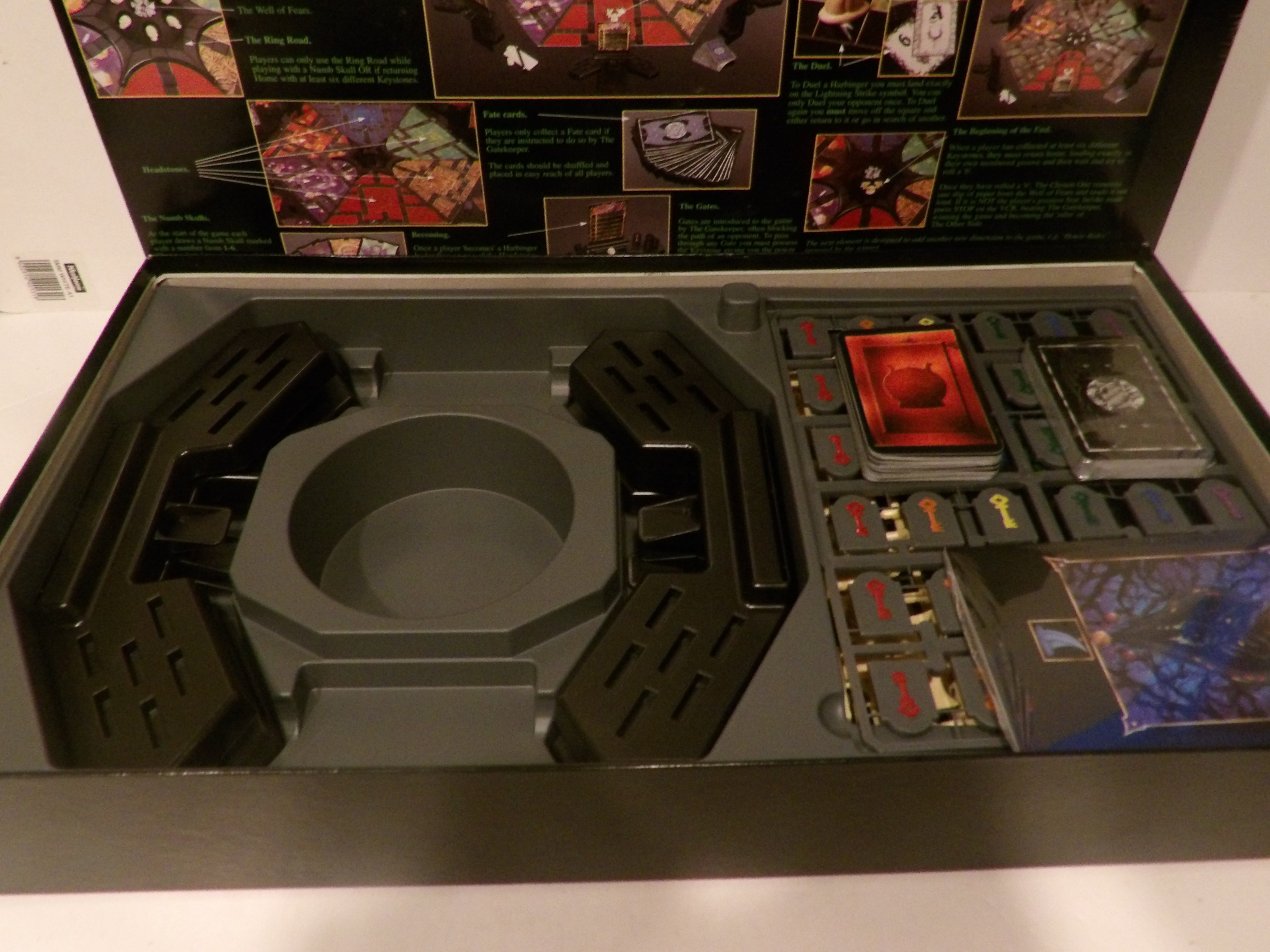Open box containing a dark gray plastic molded tray with two black wing-like pieces and a set of small game components, cards, and a blue illustrated booklet on a white background.