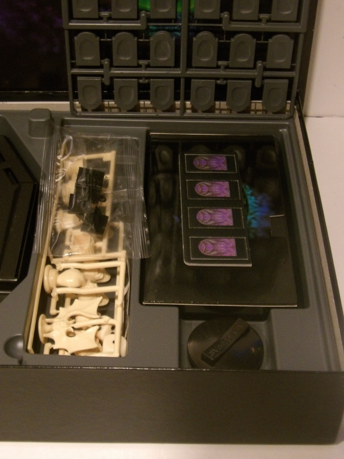 An open dark gray storage or display case containing multiple compartments with miniature accessories, including a clear plastic bag with small white and black pieces, several cards or inserts with purple graphics, and a round black item.