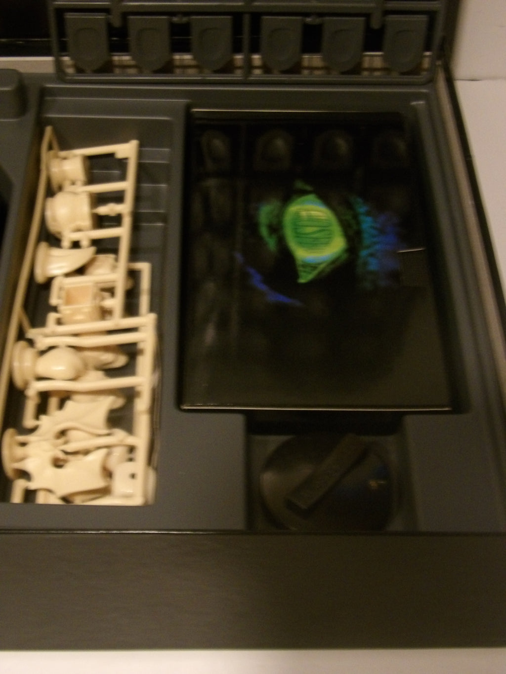 A close-up of a dark gray or black storage box or tray containing a beige plastic miniature figure set on the left and a glossy black insert with a green circular emblem in the center.