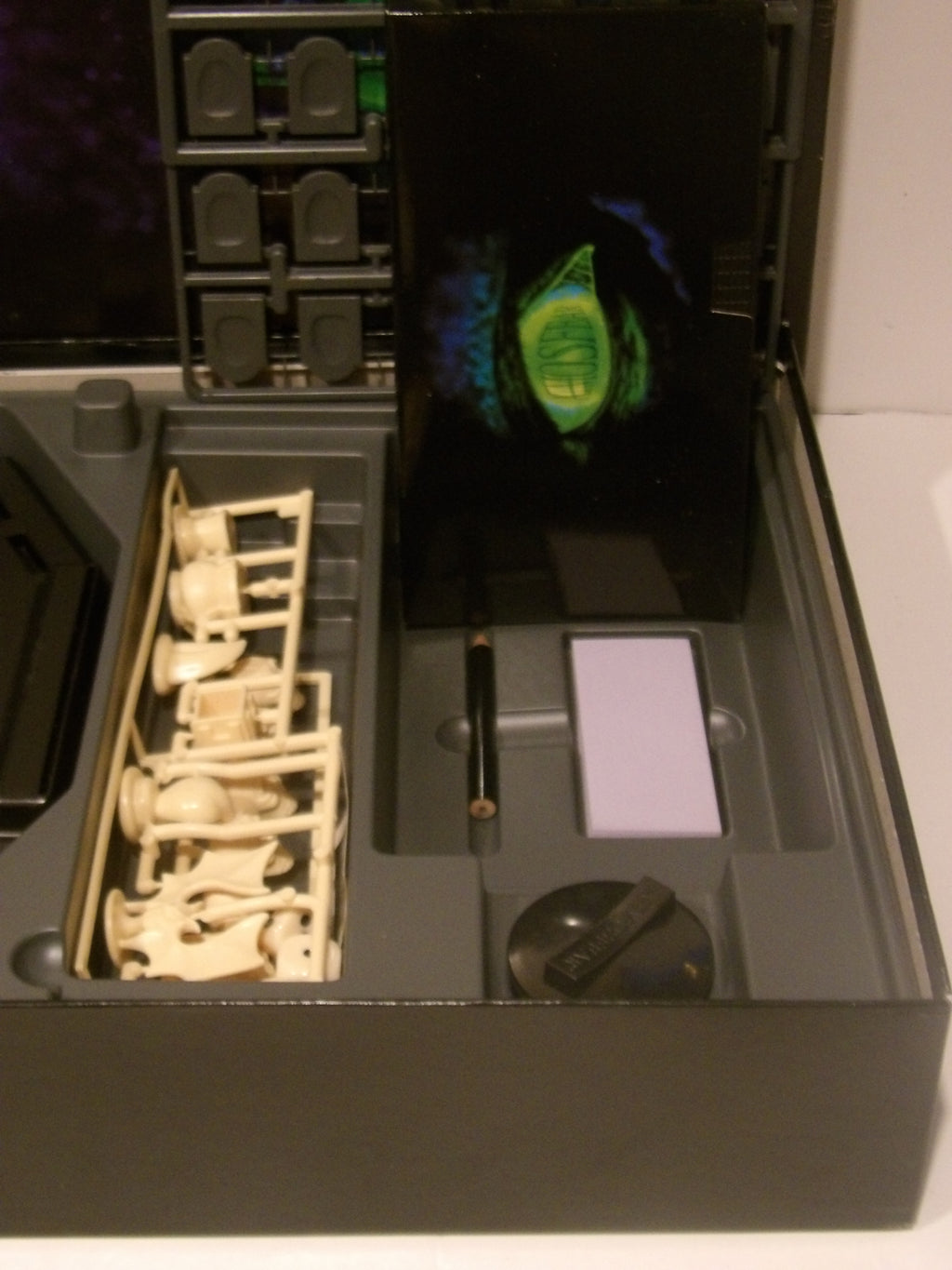 An open dark gray storage or display case showing various accessories and components, including a tray of small beige figurines or parts, a black rectangular item with a green graphic insert, a pen, a small light purple square, and a round black object.