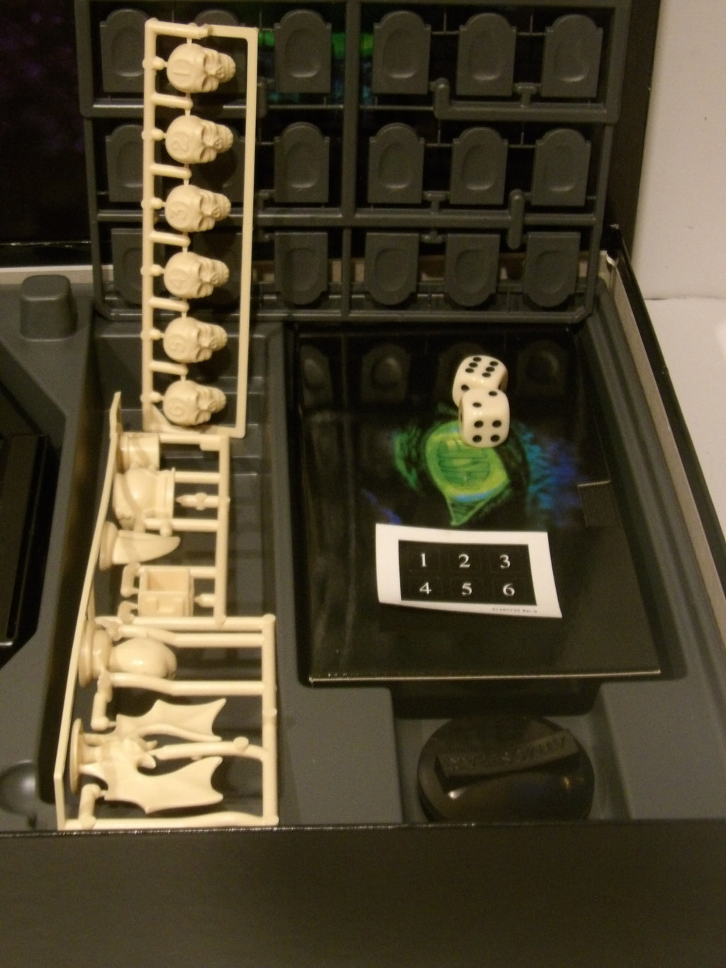 A miniature tabletop gaming accessory set displayed inside a black plastic storage box. The set includes a vertical bone rack holding small white bone-like pieces, a vertical bone rack on the left, a dice cup with a white die, a green-and-blue card, and a white numbering card showing 1 through 6.