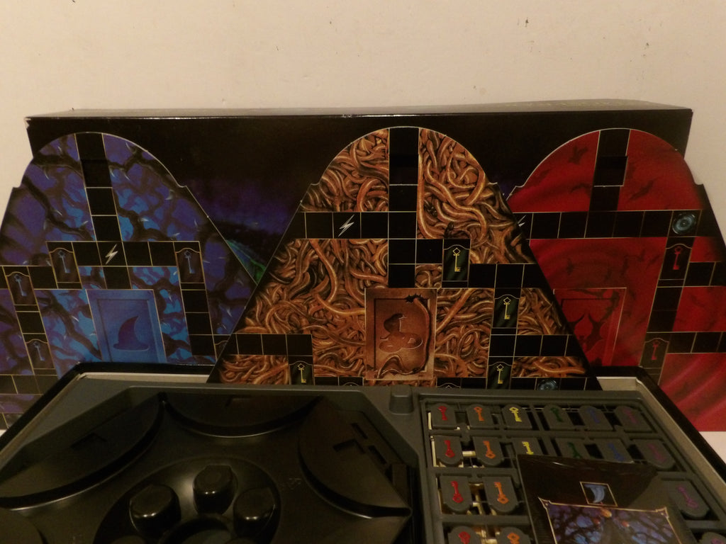 A board game set with a black control panel in the foreground and three colorful illustrated arch-shaped backdrops or panels behind it, featuring blue, brown, and red designs on a plain wall background.