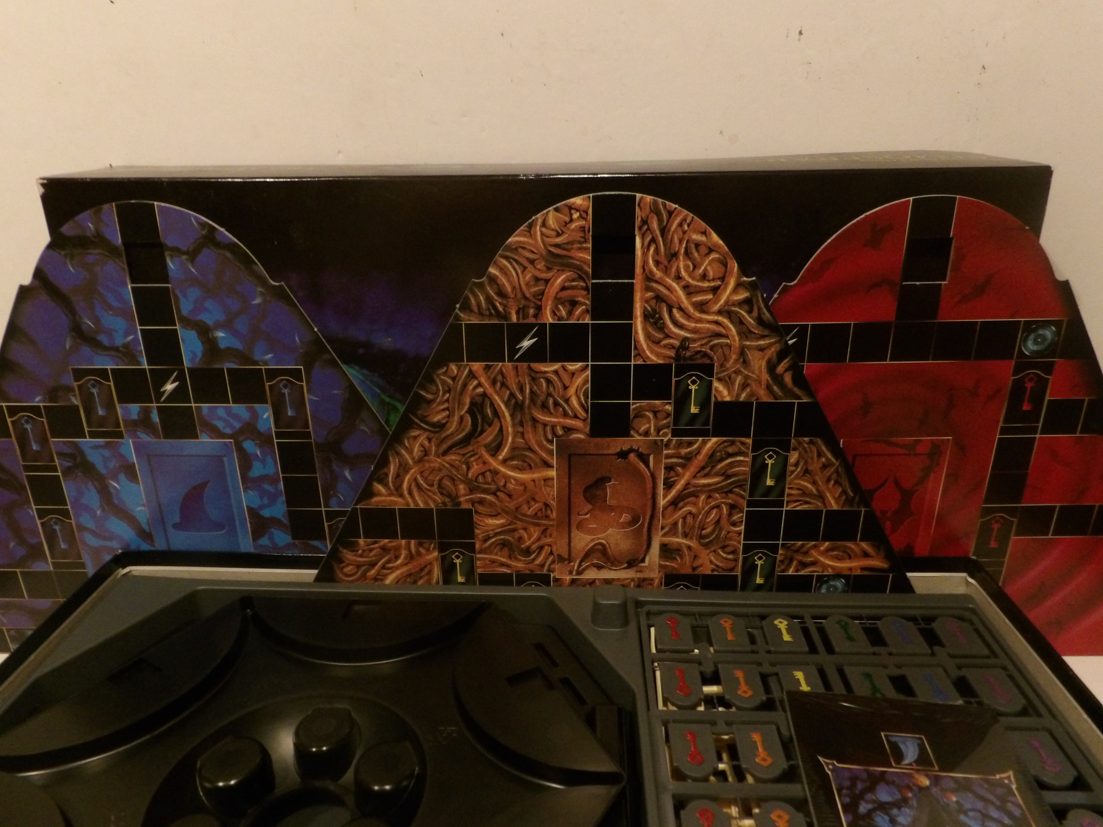 A board game set with a black control panel in the foreground and three colorful illustrated arch-shaped backdrops or panels behind it, featuring blue, brown, and red designs on a plain wall background.