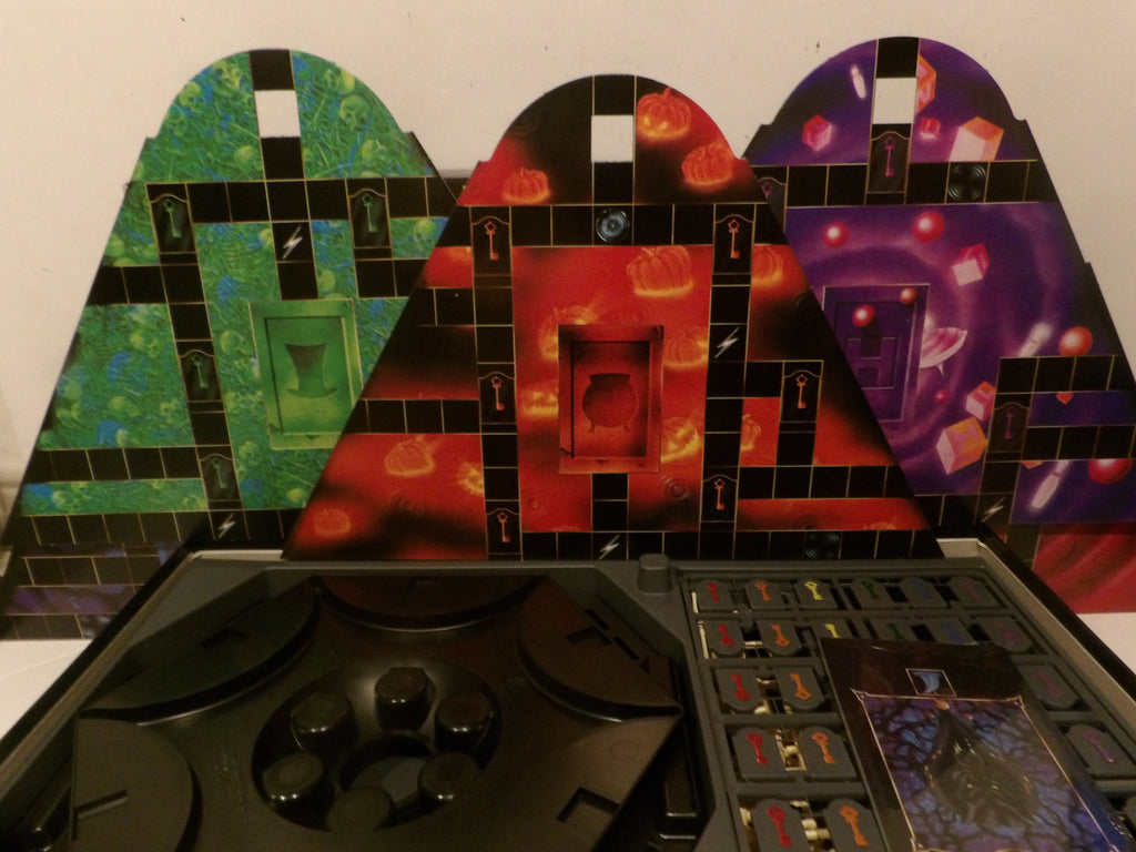 A board game set with three colorful triangular terrain panels featuring green, red, and purple fantasy designs, displayed behind a black game board with cards and tokens.