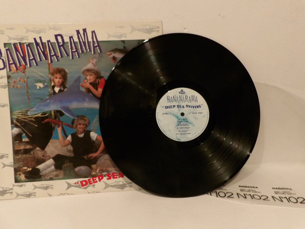 A vinyl record partially covering its printed album cover, photographed on a light surface. The black record is labeled BANANARAMA "Deep Sea" and the cover art shows people and a shark.