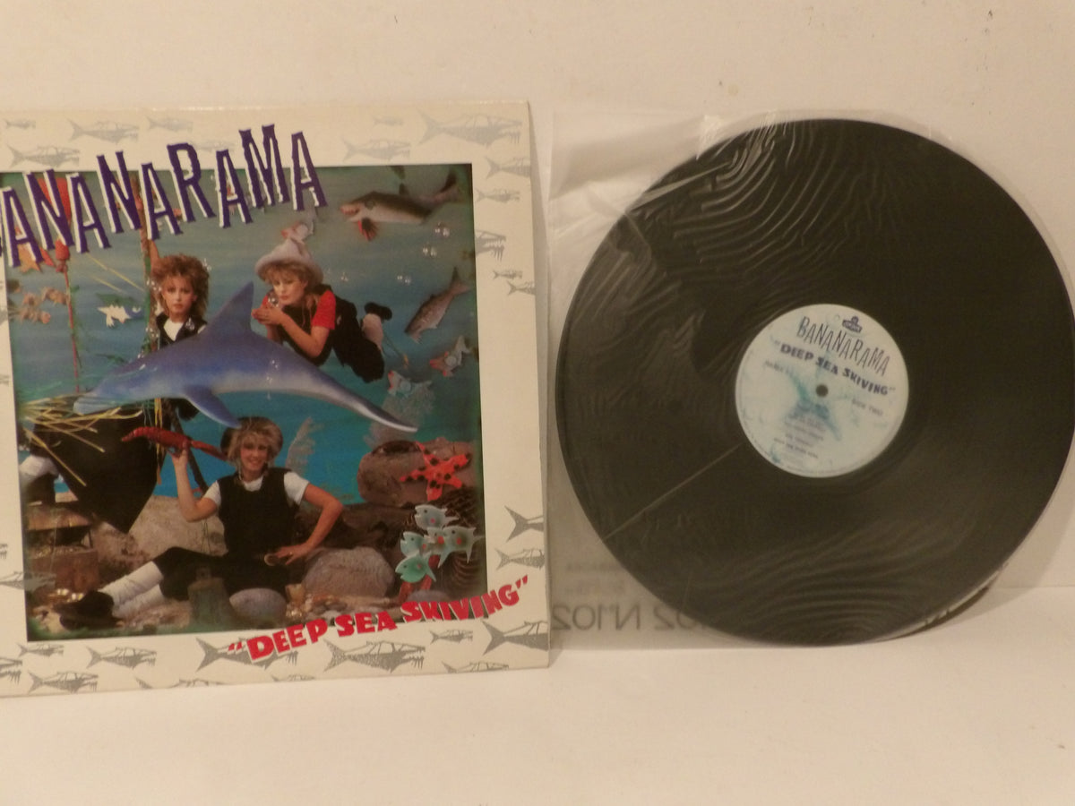 A vinyl record and its album cover for Ananarama's 'Deep Sea Shining' are displayed on a light-colored surface. The black vinyl record is partially inside a clear protective sleeve, and the illustrated cover features a colorful underwater scene with a dolphin and the band members.