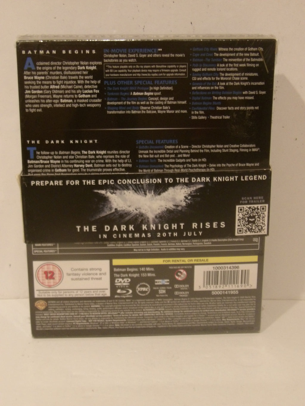 Back cover of The Dark Knight Rises DVD box set, showing release information, synopsis text, barcode, and age rating on a light background.