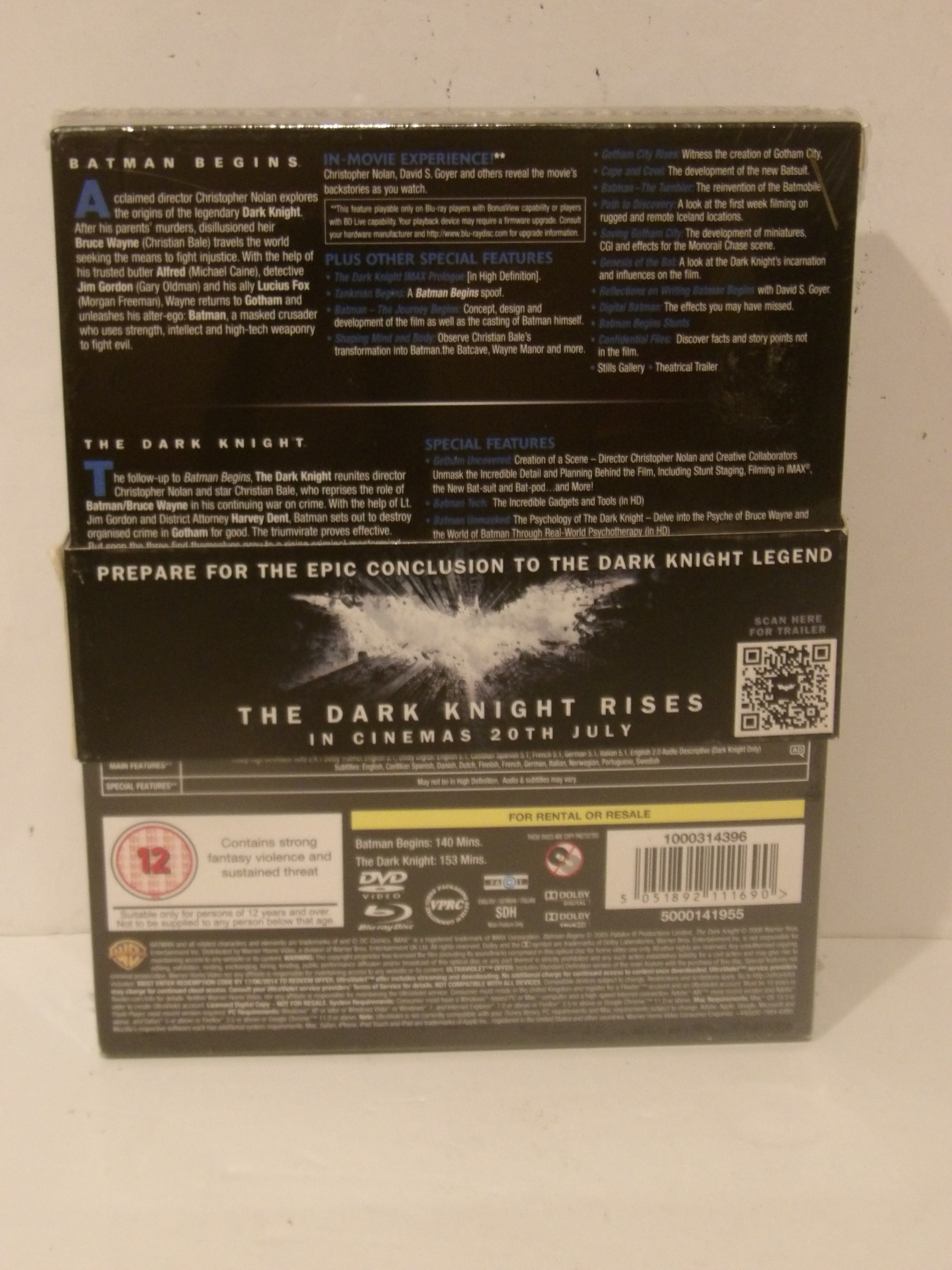 Back cover of The Dark Knight Rises DVD box set, showing release information, synopsis text, barcode, and age rating on a light background.