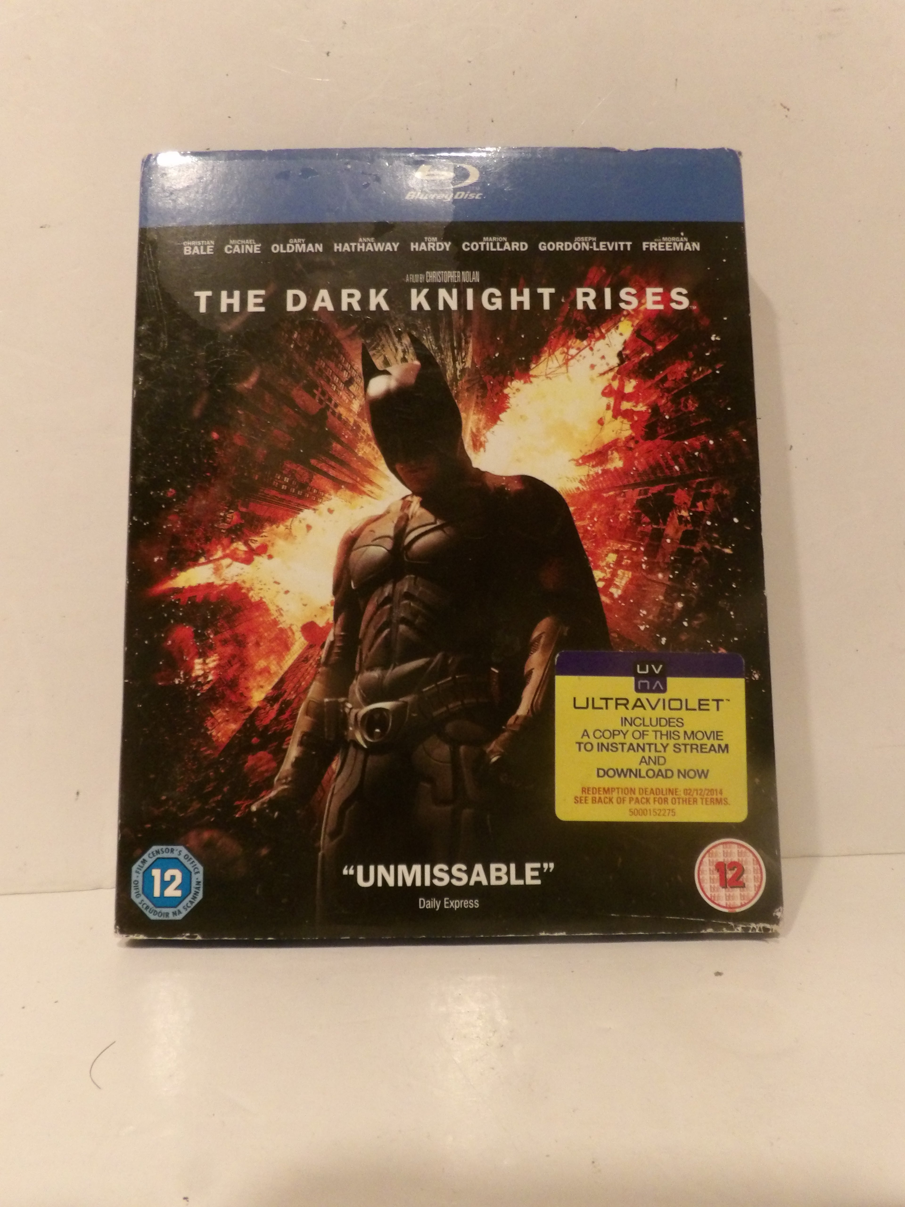 The Dark Knight Rises Blu-ray movie case on a light beige surface. The cover features Batman in front of a fiery explosion, with a yellow Ultraviolet sticker and a blue Blu-ray banner at the top.