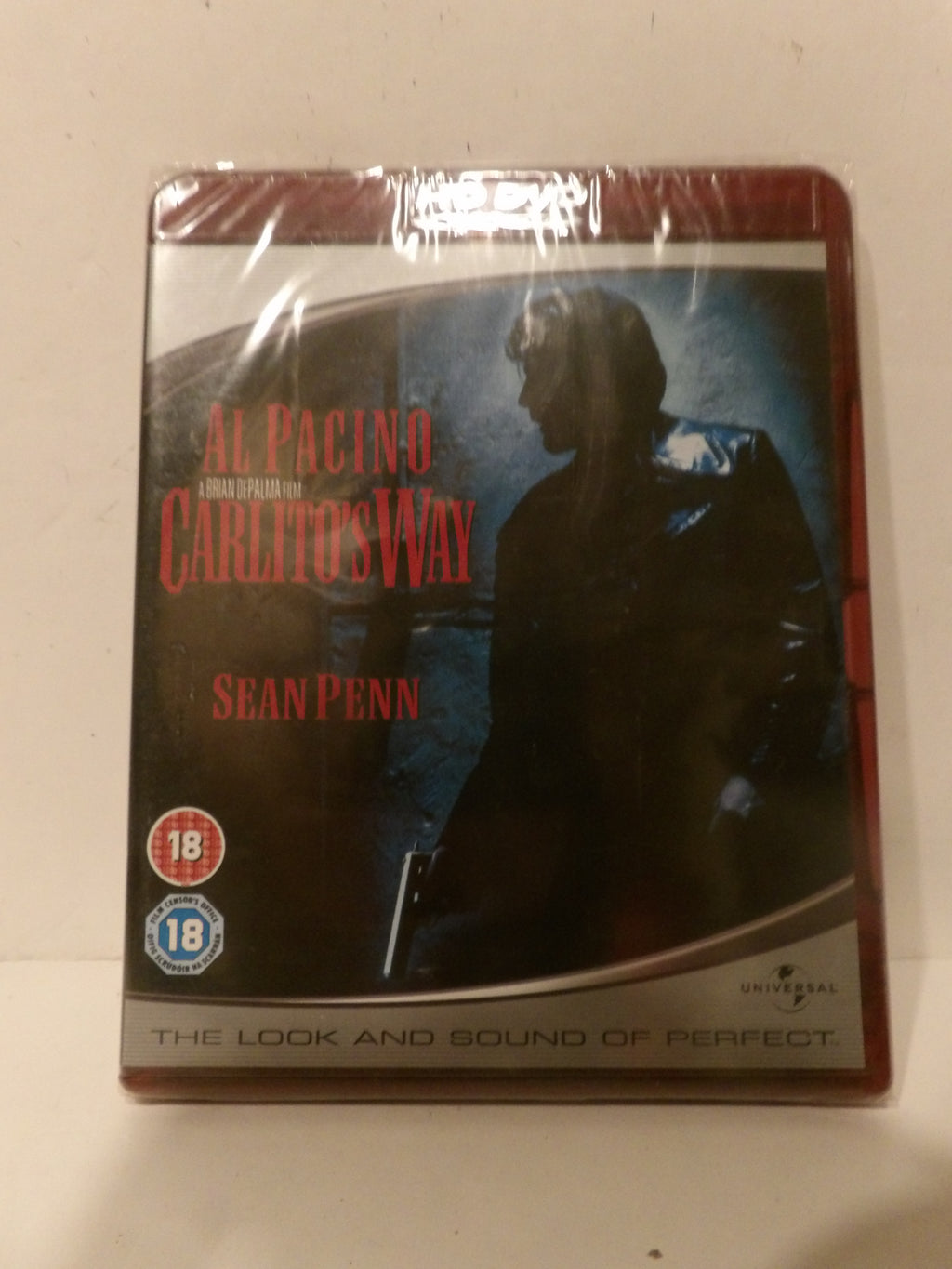 A sealed DVD case of Al Pacino's 'The Godfather: Carlito's Way' featuring Sean Penn, photographed on a light beige surface. The case is wrapped in protective plastic and shows a red spine and a dark cover with red title text.