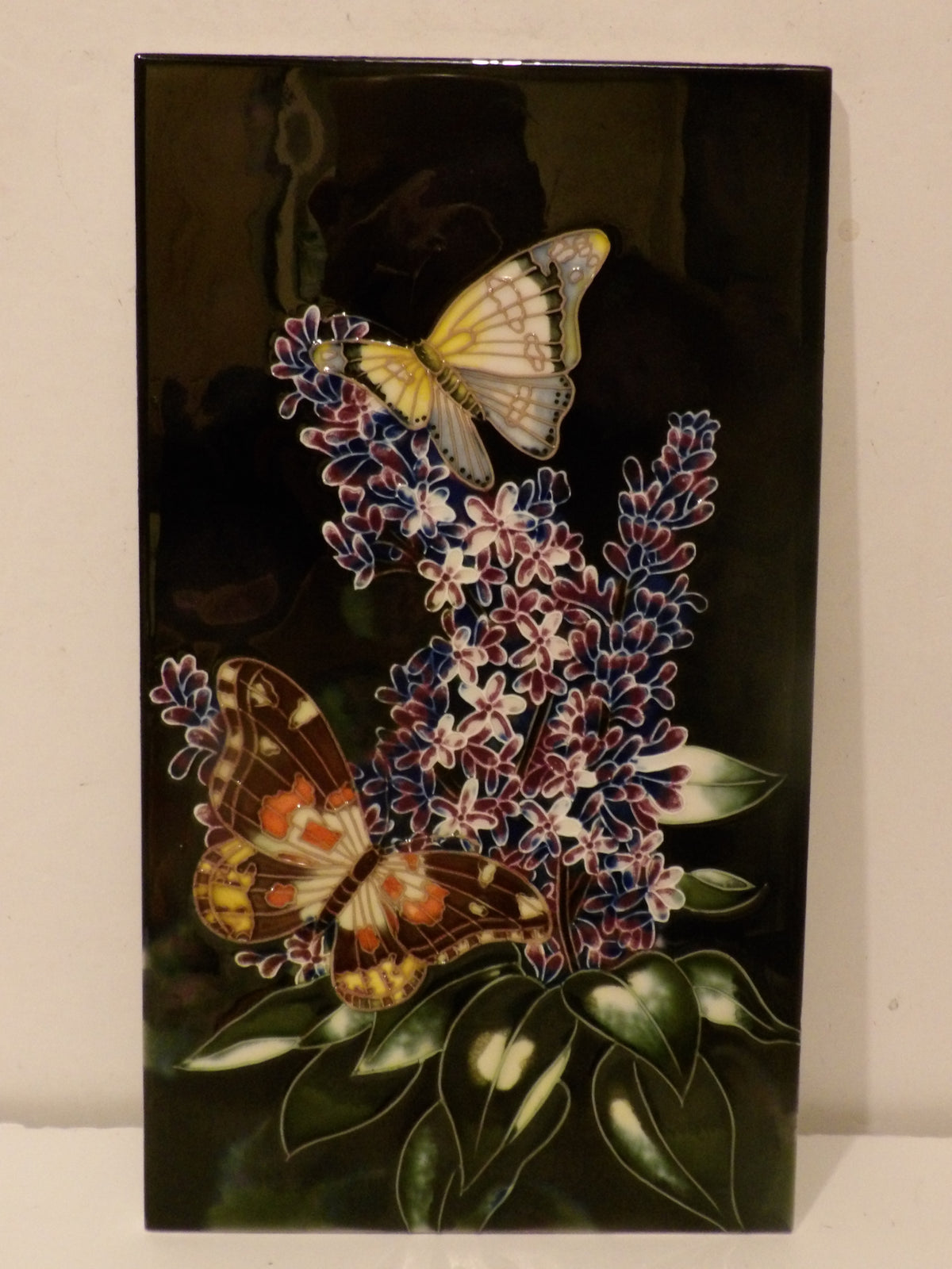 A rectangular glossy black decorative tile featuring a detailed illustration of two butterflies perched on purple and white hyacinth flower clusters with green leaves.