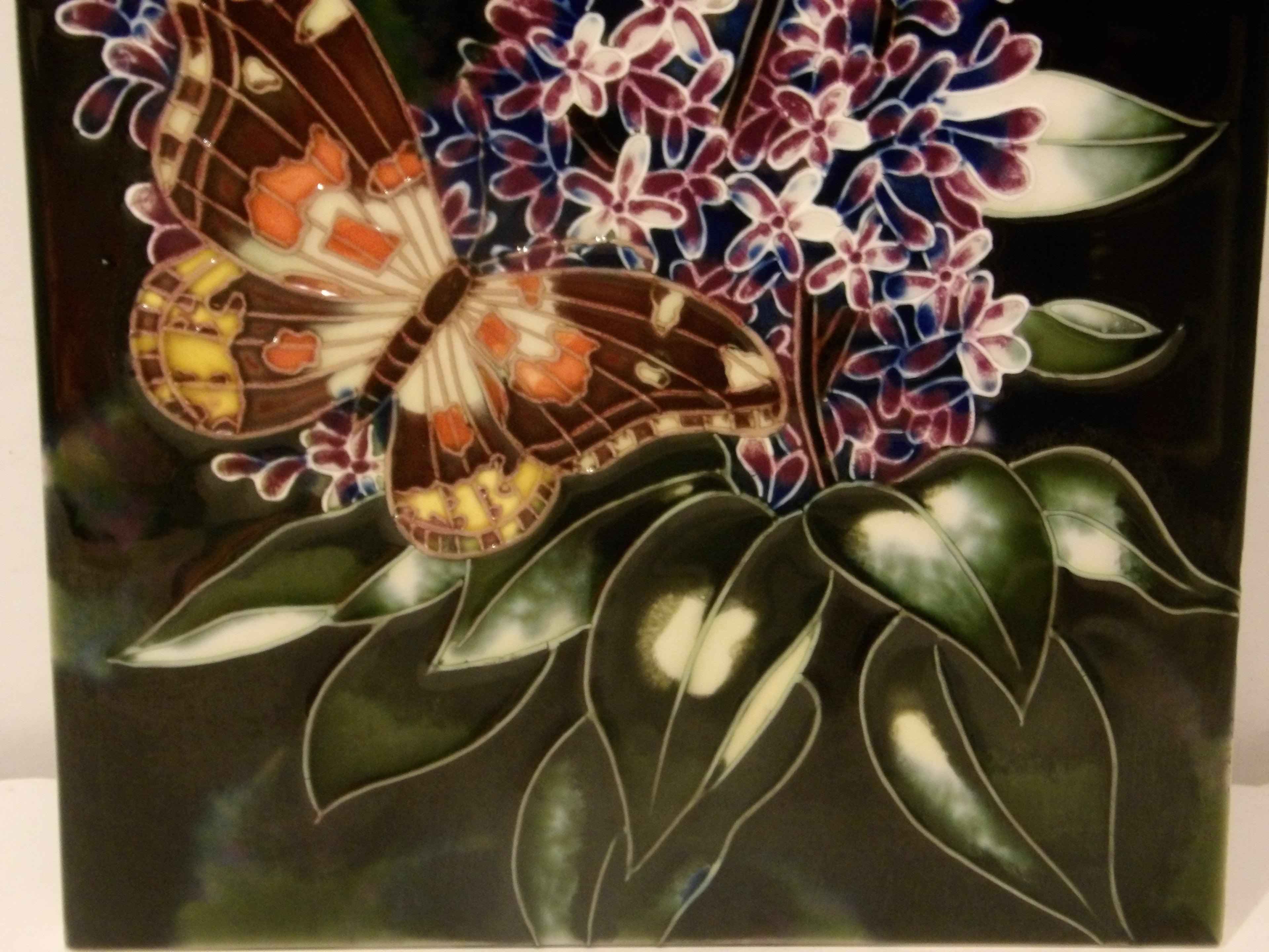 A square ceramic tile featuring a detailed illustration of a brown and orange butterfly perched on purple and white flowers with dark green leaves against a black background.