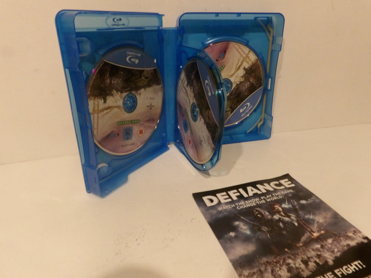 A blue Blu-ray case standing open with two discs visible, alongside a printed insert or booklet labeled 'DEFIANCE' on a plain light surface.