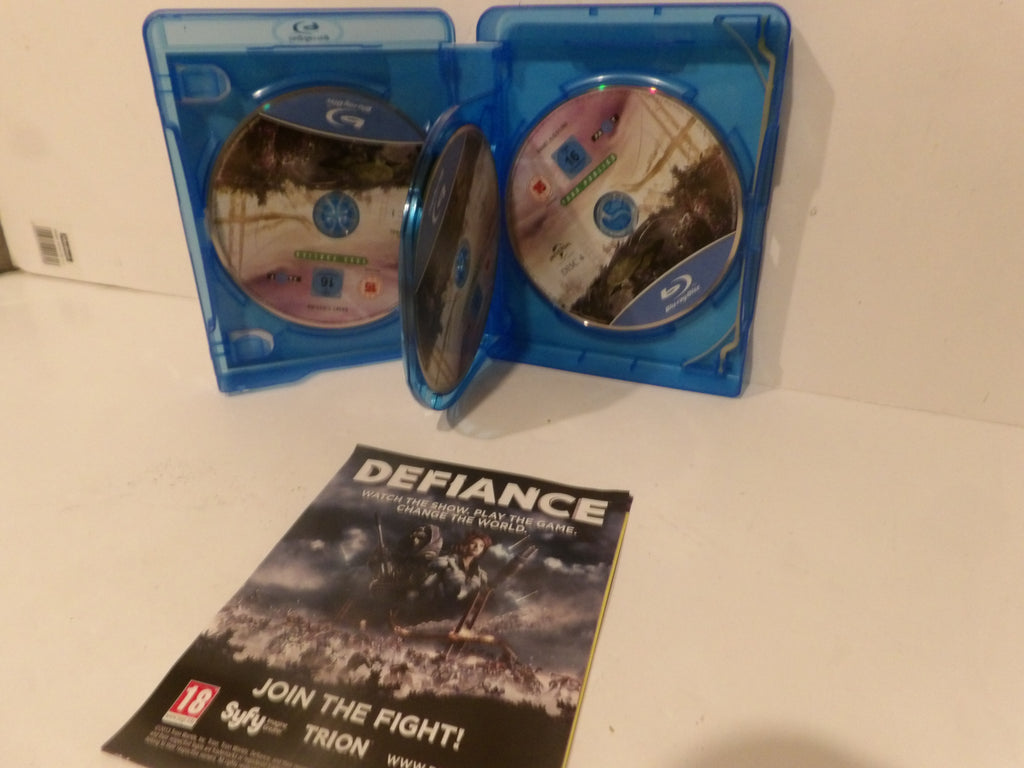 Defiance video game Blu-ray case set with two blue plastic cases, one open and displaying the discs, a second disc leaning in front, and a printed insert card on a white surface.