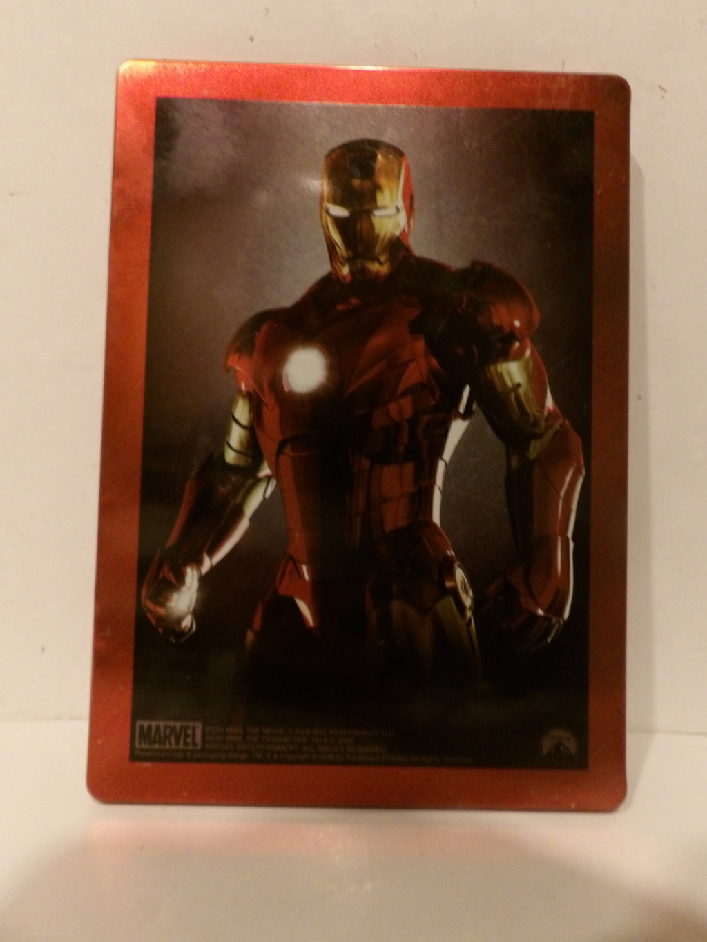 A rectangular collectible card featuring Iron Man in a dark, dramatic pose with a metallic red border, photographed against a plain light background.