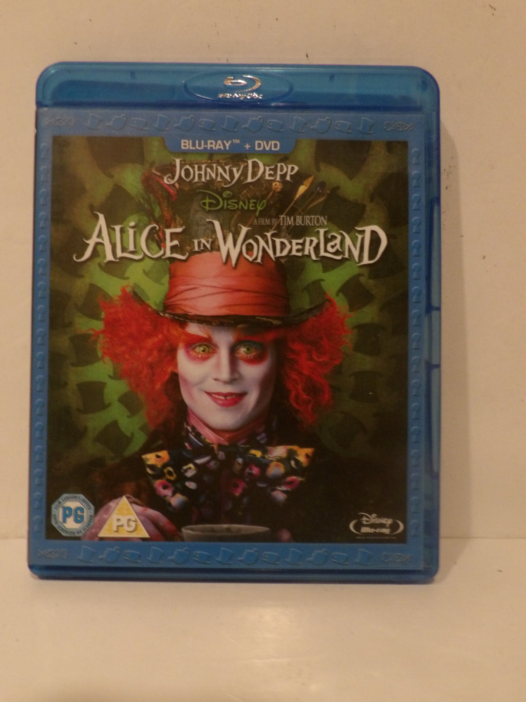 A Blu-ray and DVD combo pack of Disney's 'Alice in Wonderland' featuring Johnny Depp as the Mad Hatter, shown in its blue plastic case on a light beige surface.