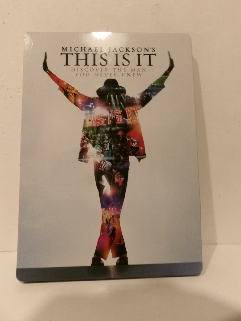 Michael Jackson's 'This Is It' steelbook Blu-ray case standing upright against a light beige wall. The cover features a silver-gray background with a colorful silhouette of Michael Jackson in a rainbow jacket and hat, with the title 'THIS IS IT' in bold black letters at the top.