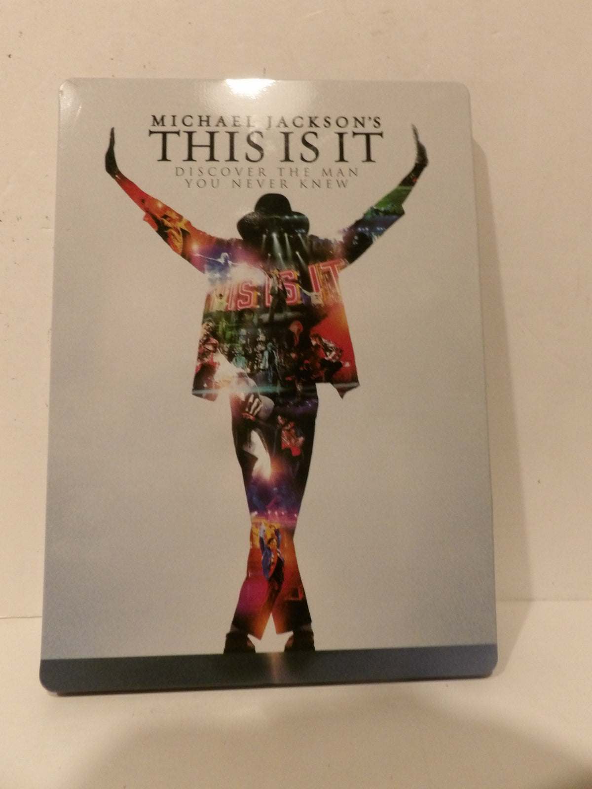 Michael Jackson's 'This Is It' steelbook Blu-ray case standing upright against a light beige wall. The cover features a silver-gray background with a colorful silhouette of Michael Jackson in a rainbow jacket and hat, with the title 'THIS IS IT' in bold black letters at the top.