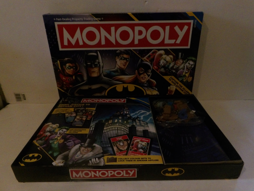 Batman-themed Monopoly board game set with the main game box standing upright behind an open tray displaying cards and play money on a light-colored surface.