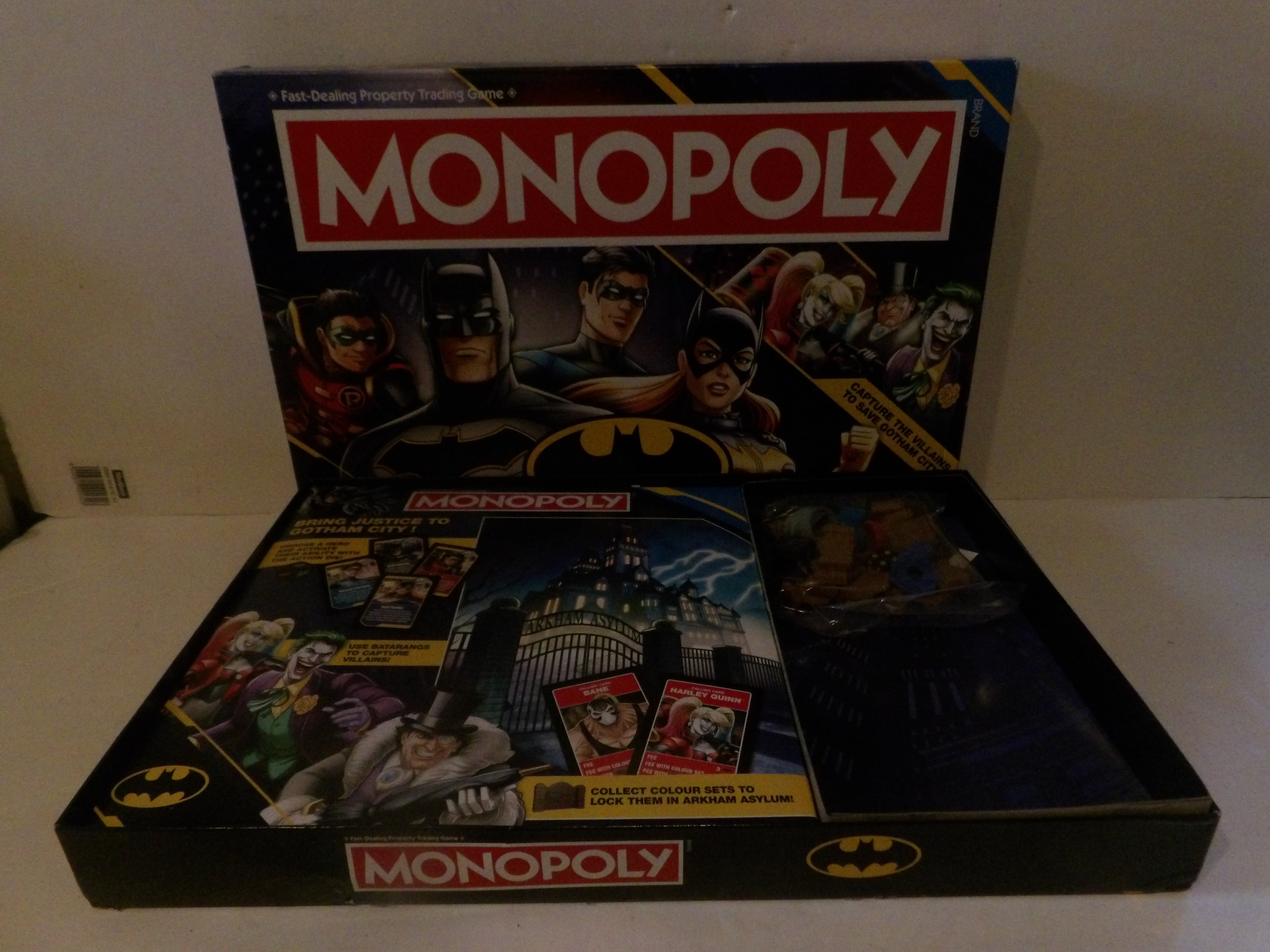 Batman-themed Monopoly board game set with the main game box standing upright behind an open tray displaying cards and play money on a light-colored surface.