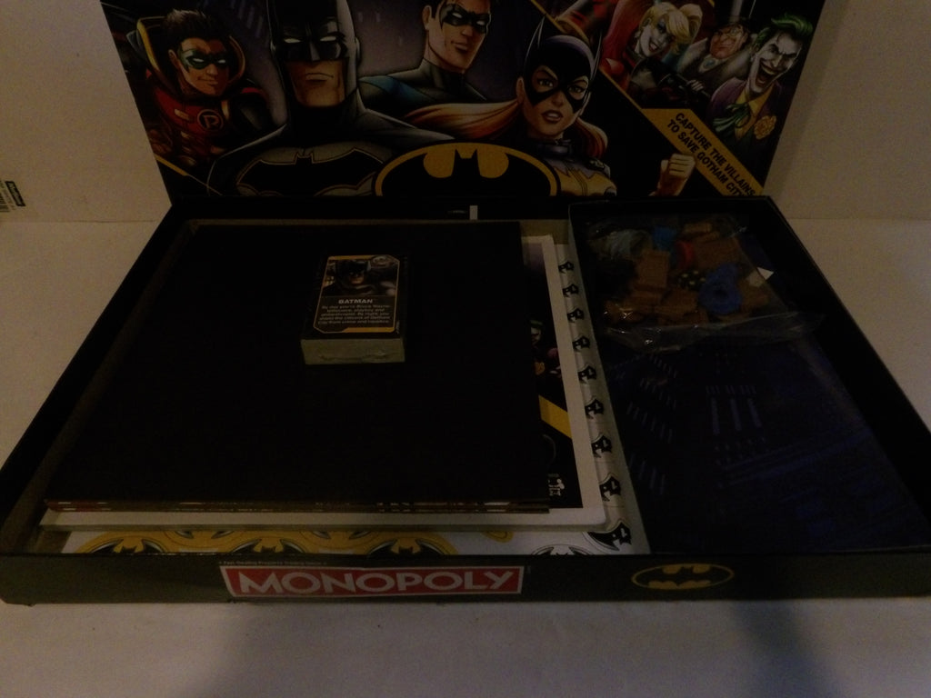 A Batman-themed Monopoly board game set opened to show the interior contents, including the game board, play money, cards, and a small booklet, with Batman and other DC characters printed on the inside lid.