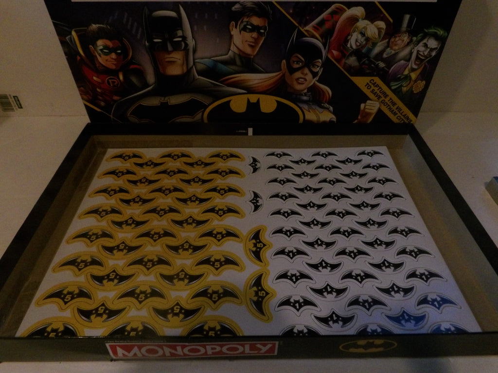 A Monopoly board game set featuring Batman-themed play money inside a black case, with illustrated Batman character artwork on the inside lid.