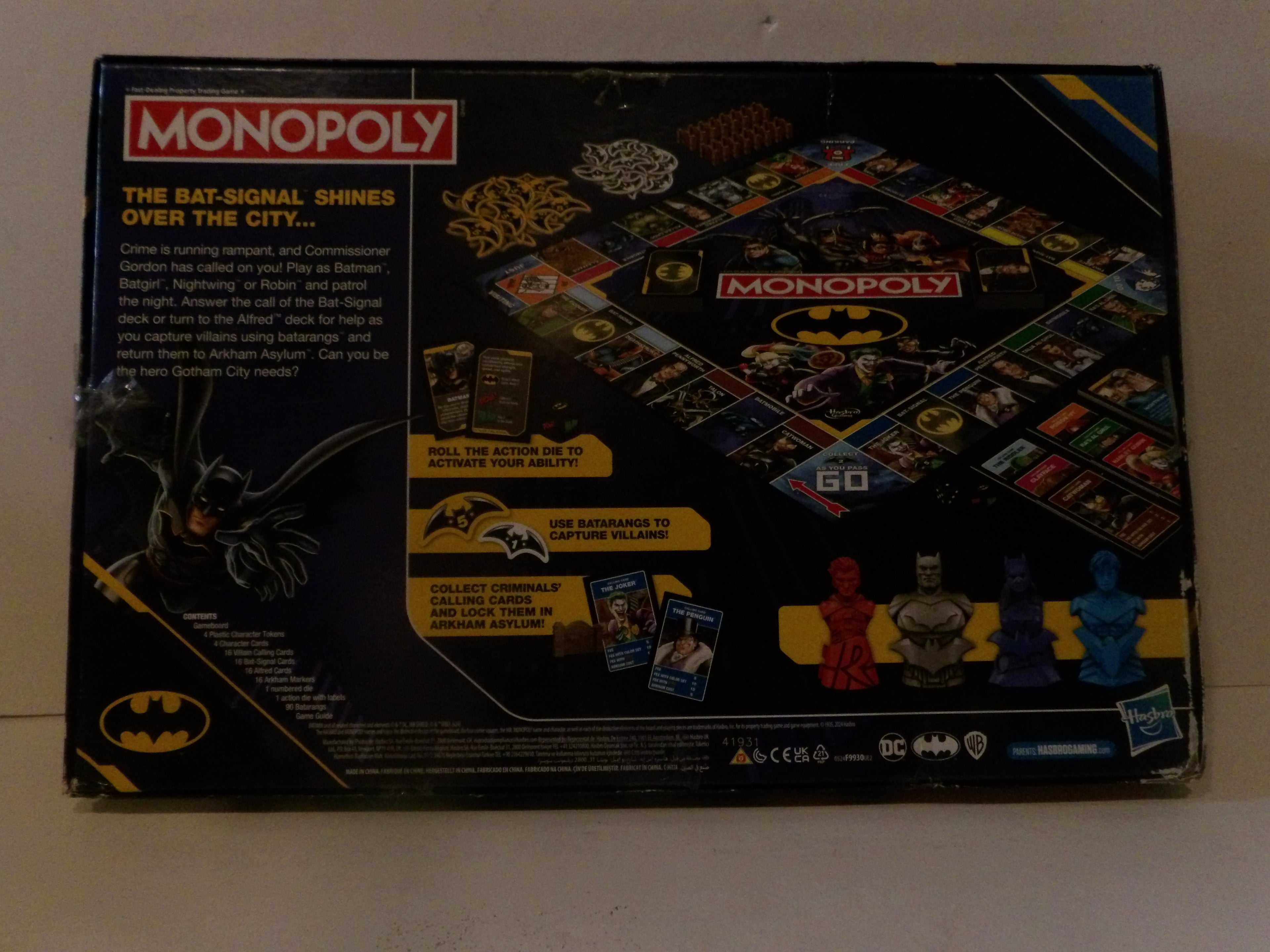 The back of a vintage Monopoly board game box featuring Batman-themed artwork, including the Batman logo, a bat-shaped signal illustration, and character pawns on a light-colored floor.