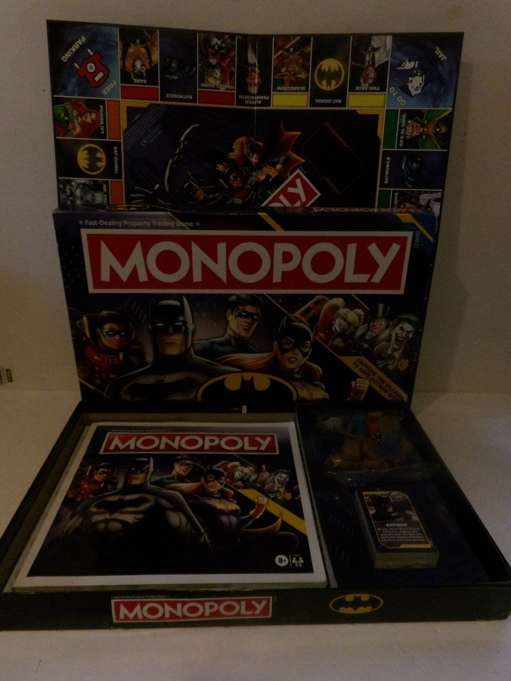 Batman-themed Monopoly board game set displayed open, showing the main game box, play money, and cards against a plain indoor background.