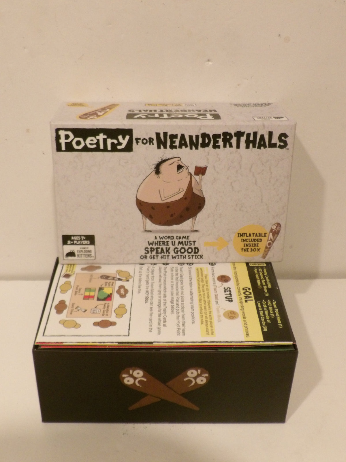 Poetry for Neanderthals board game set with its illustrated white box on top and the black game box below, photographed against a plain off-white wall.
