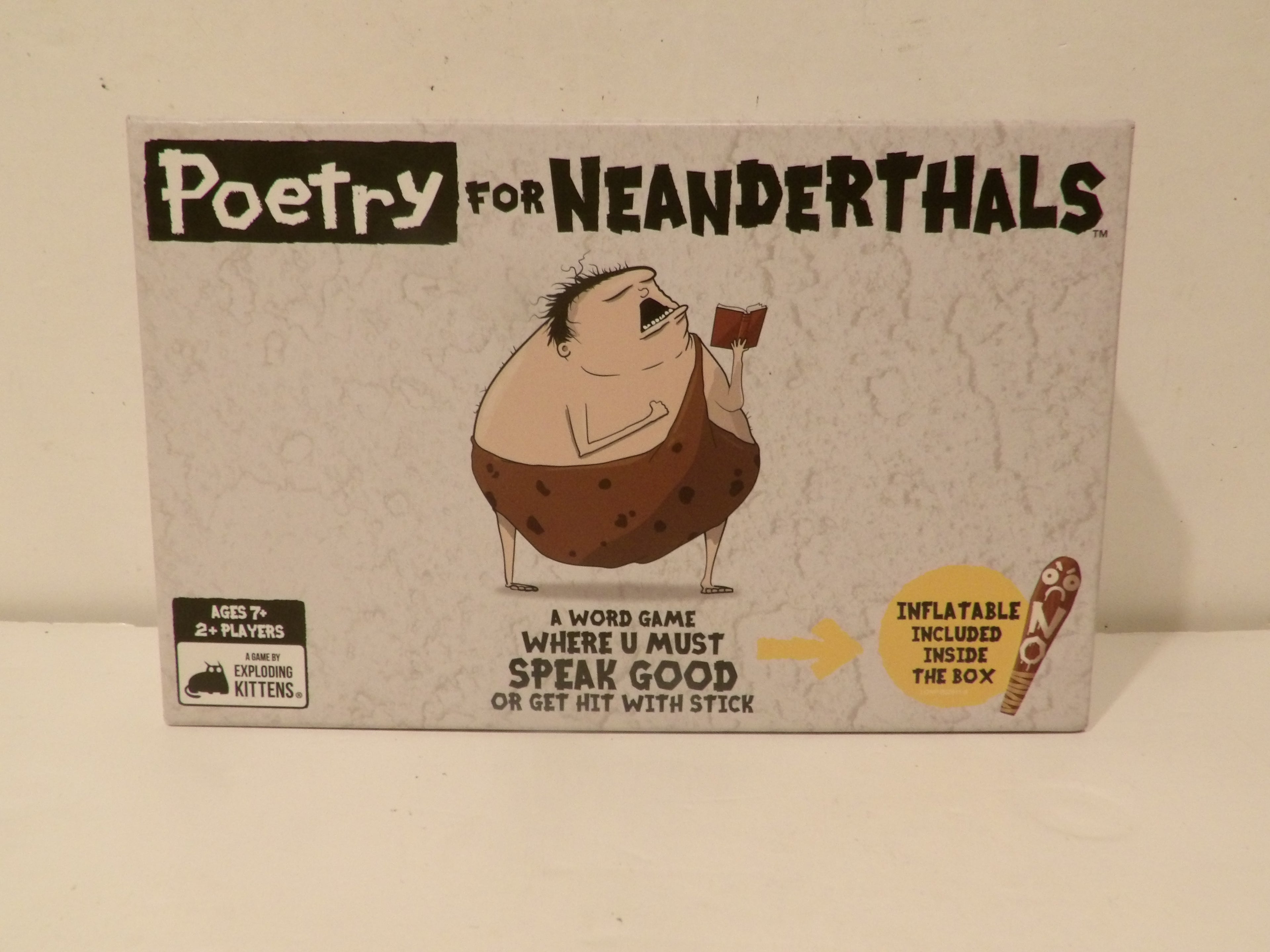 The front of the Poetry for Neanderthals board game box, featuring a cartoon illustration of a Neanderthal holding a small red flag. The box is off-white with black and brown graphics and text, photographed against a plain beige background.