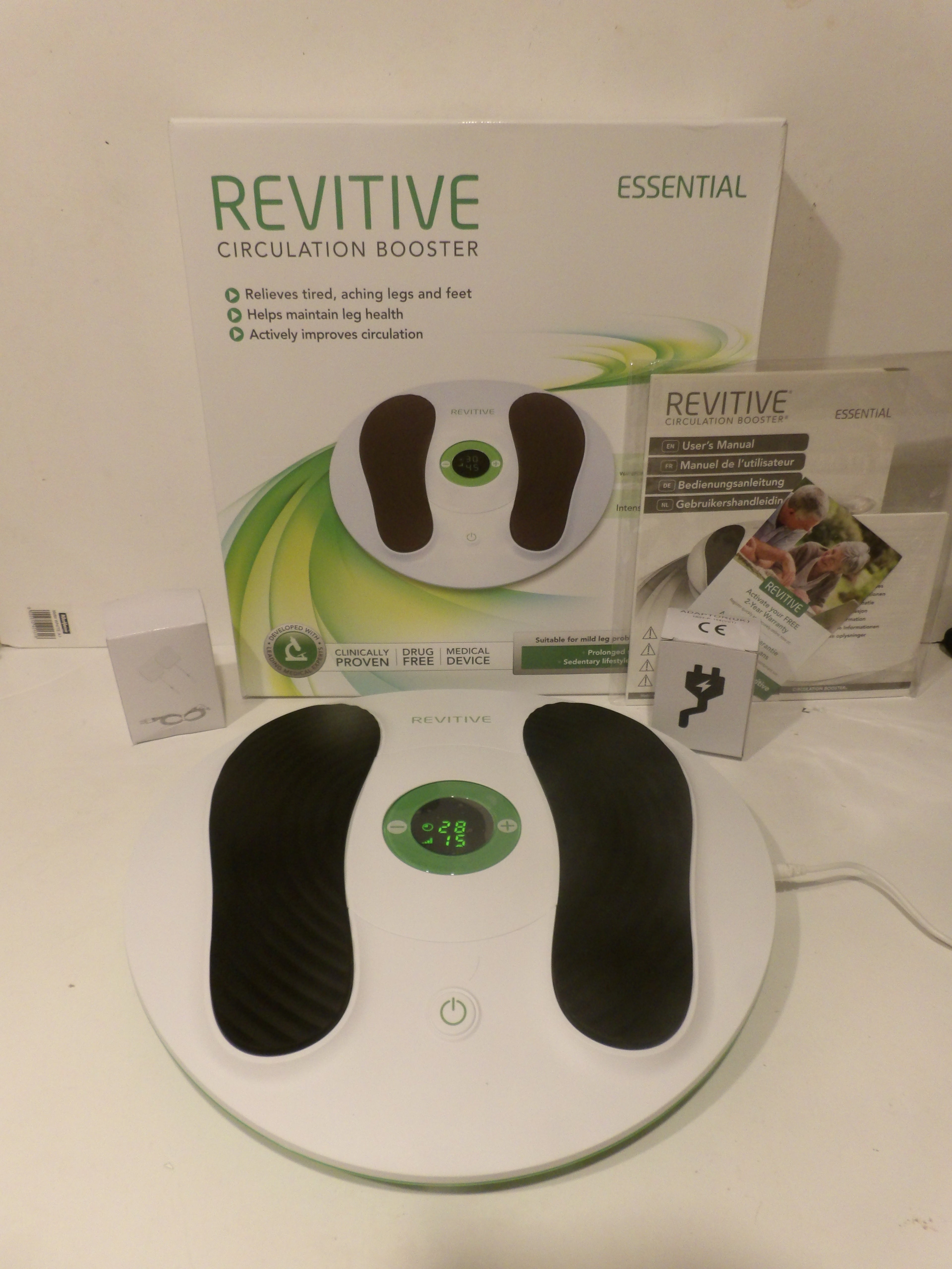 A Revitive Essential Circulation Booster foot massager shown with its retail box, manual, and accessories on a white surface against a white wall.