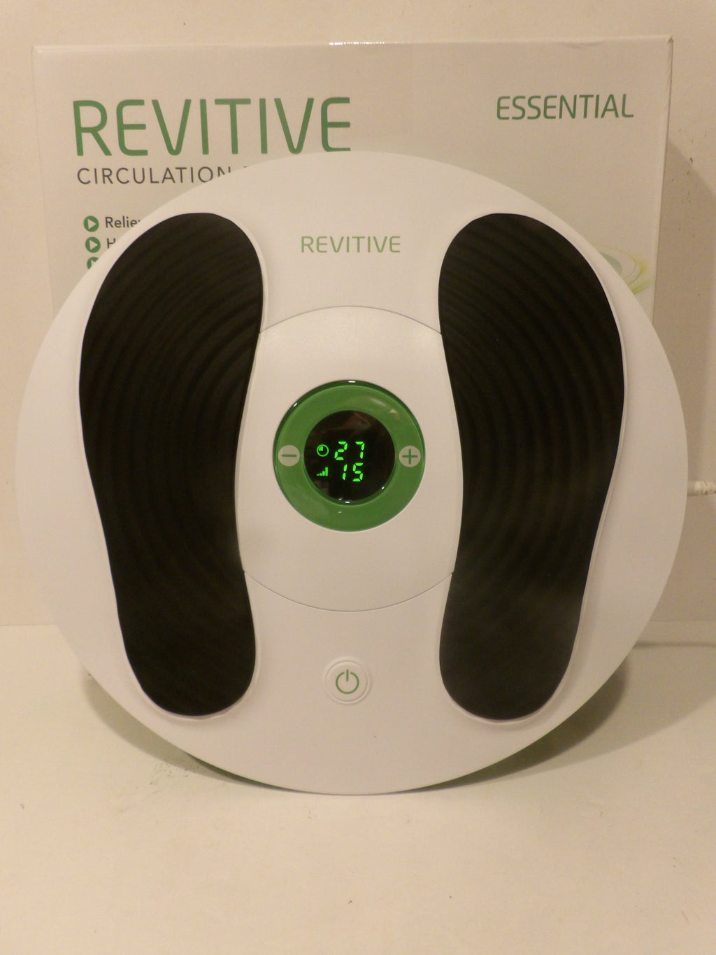 A white REVITIVE Essential Circulation Massager shown with its white retail box in the background. The device features two black textured footrests and a central green digital display screen.