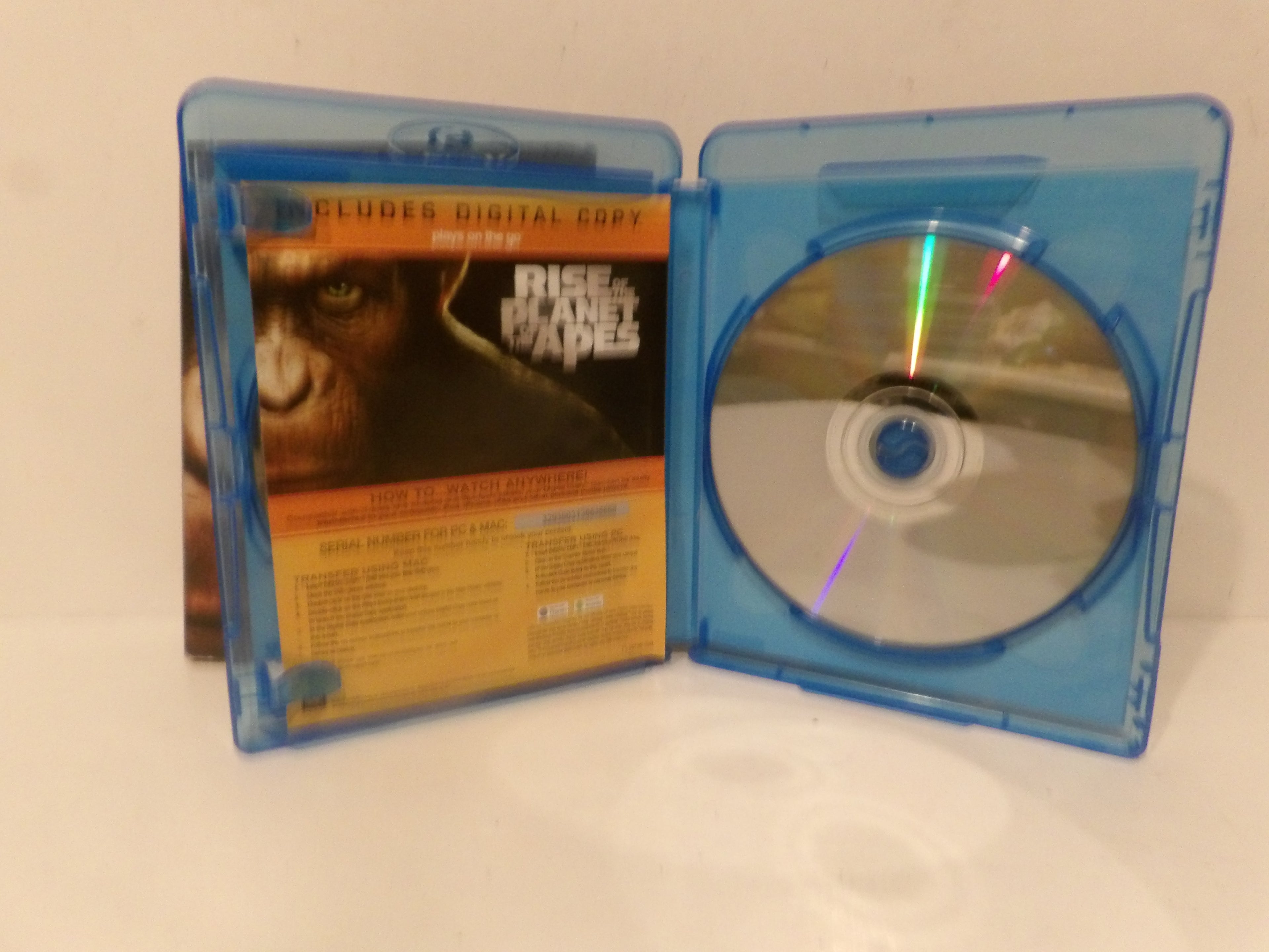 A physical copy of the movie 'Rise of the Planet of the Apes' Blu-ray in a translucent blue plastic case, shown open with the front cover insert and a silver disc inside on a white surface.