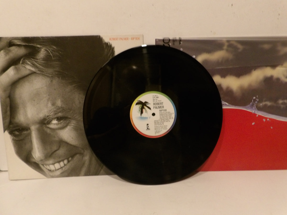 A black vinyl record with a colorful palm tree label in the center, displayed in front of its album cover. The album cover features a black-and-white close-up portrait of a smiling man on the left and a red and gray scenic design on the right.