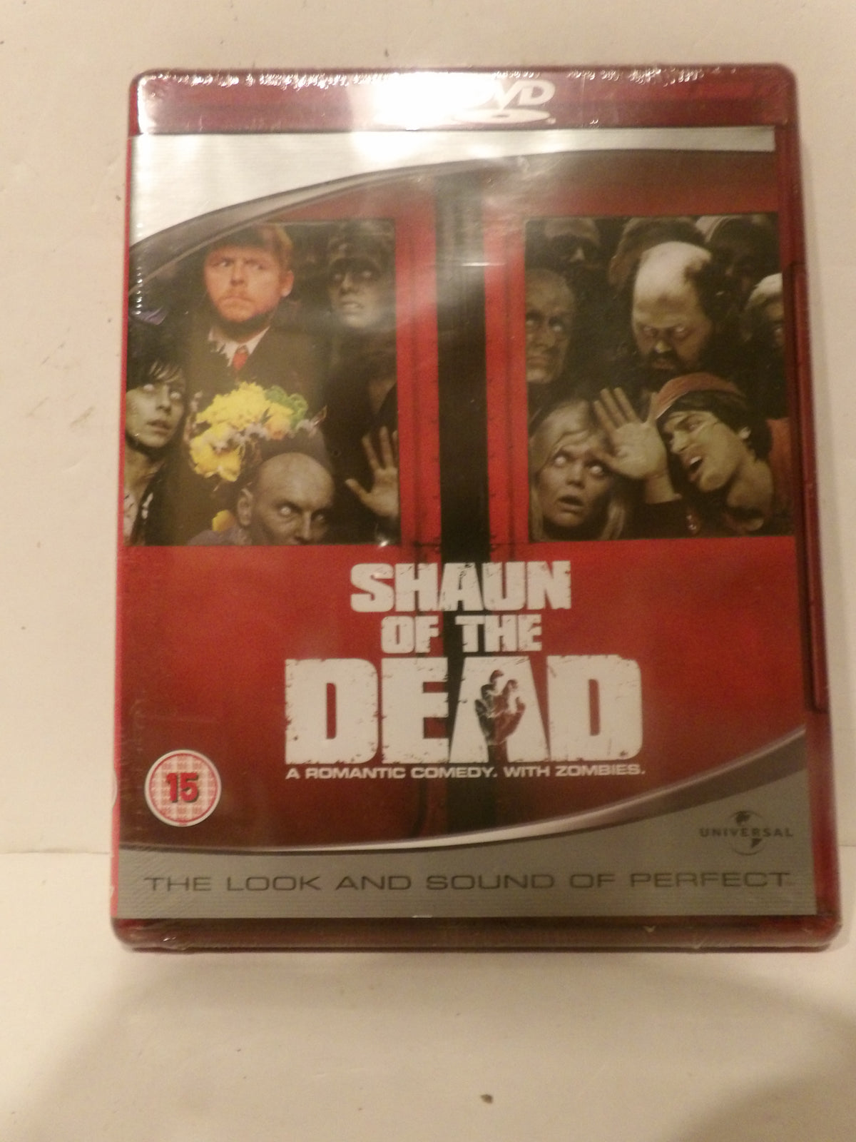 Shaun of the Dead DVD movie case, sealed in plastic shrink wrap, photographed on a light off-white surface. The cover features a red and black design with character photos and the title 'SHAUN OF THE DEAD'.