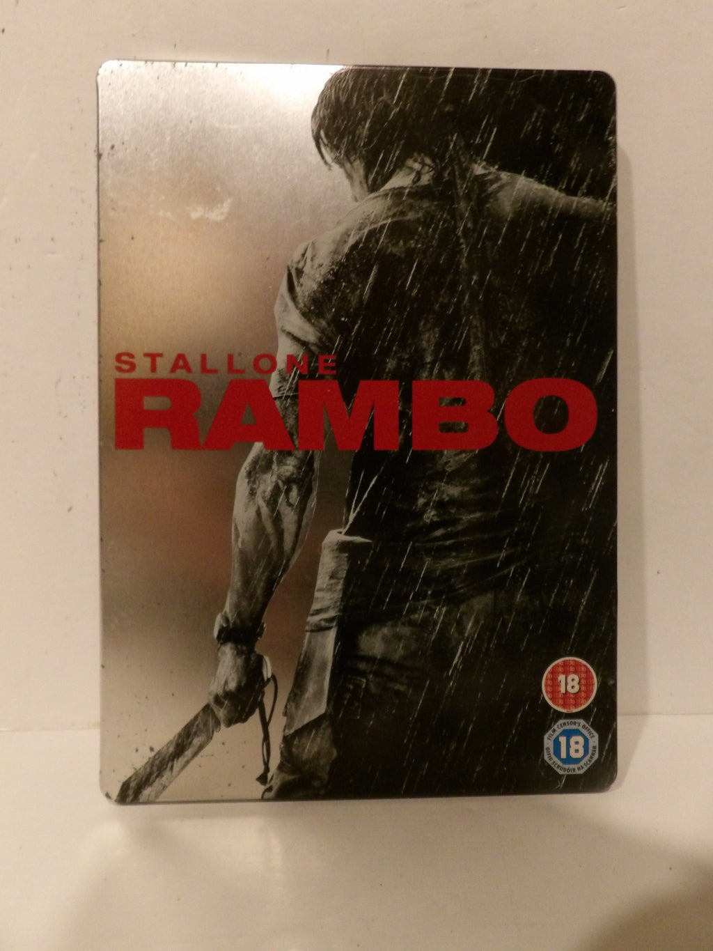 Rambo 1 film steelbook case featuring a black and white graphic of a man in the rain with bold red 'RAMBO' title text and 'STALLONE RAMBO' lettering, photographed on a light surface.