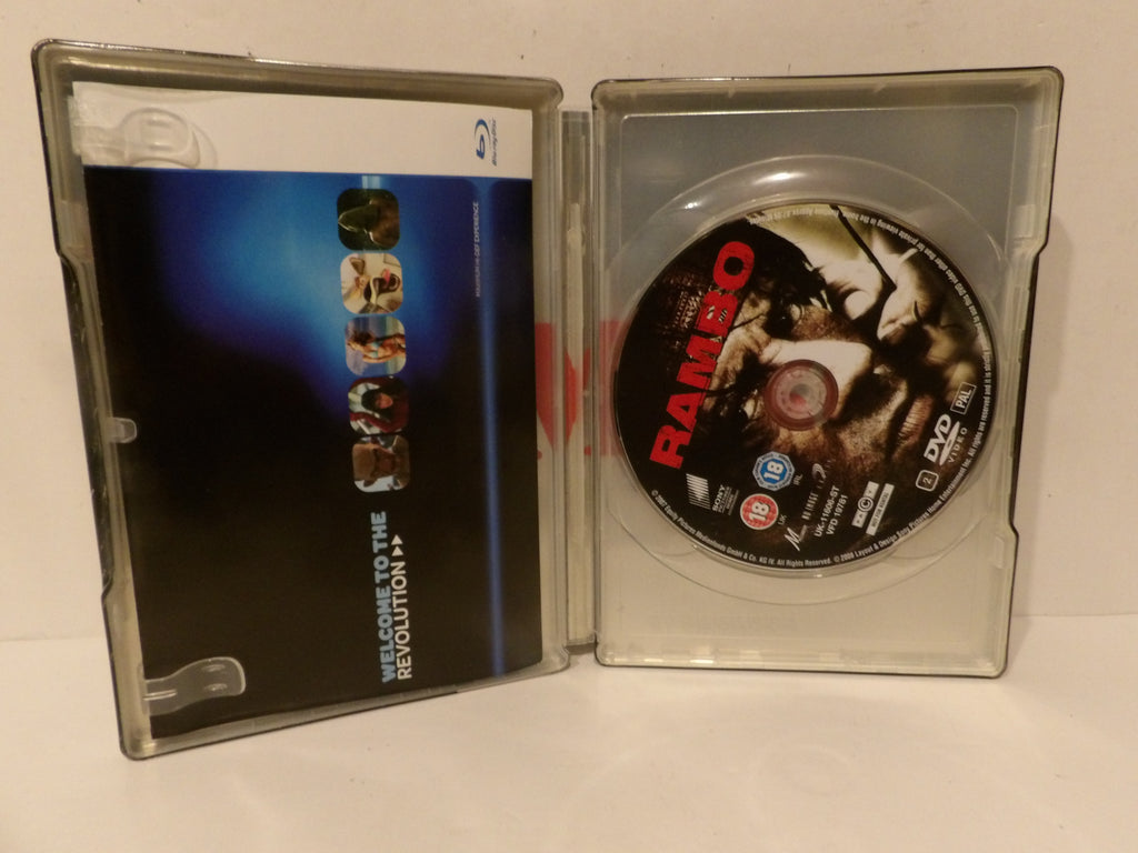 A clear plastic DVD case shown open, with the printed DVD sleeve on the left and the DVD disc inside the tray on the right. The disc features the title RAMBO in large red letters over a dark action scene.