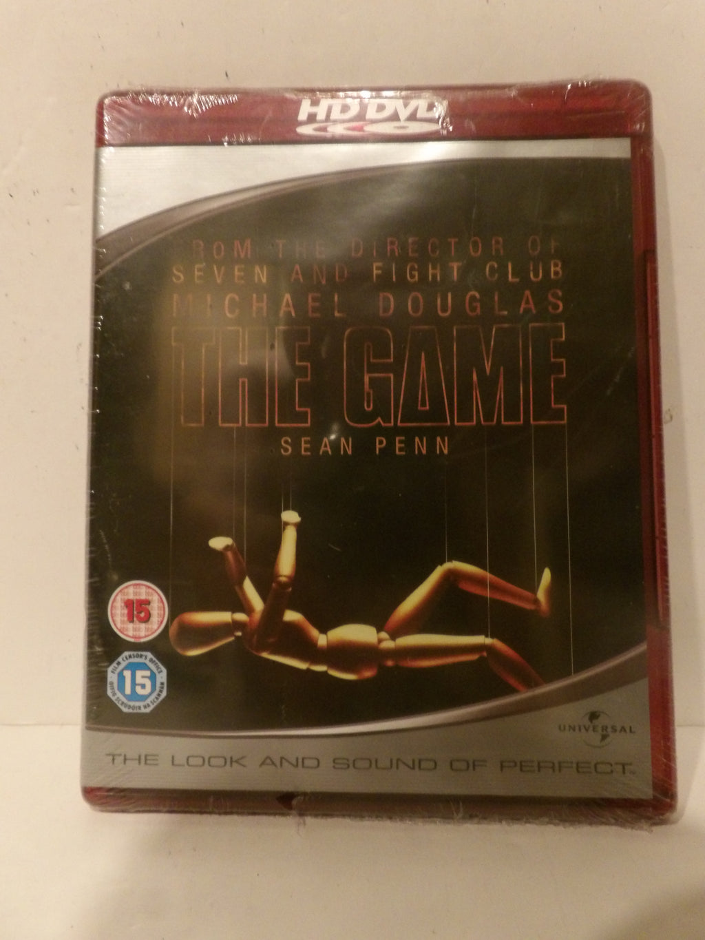 A sealed DVD box set of the movie 'The Game' starring Sean Penn, featuring a dark cover with the title in gold and a wooden mannequin figure, photographed on a light surface.