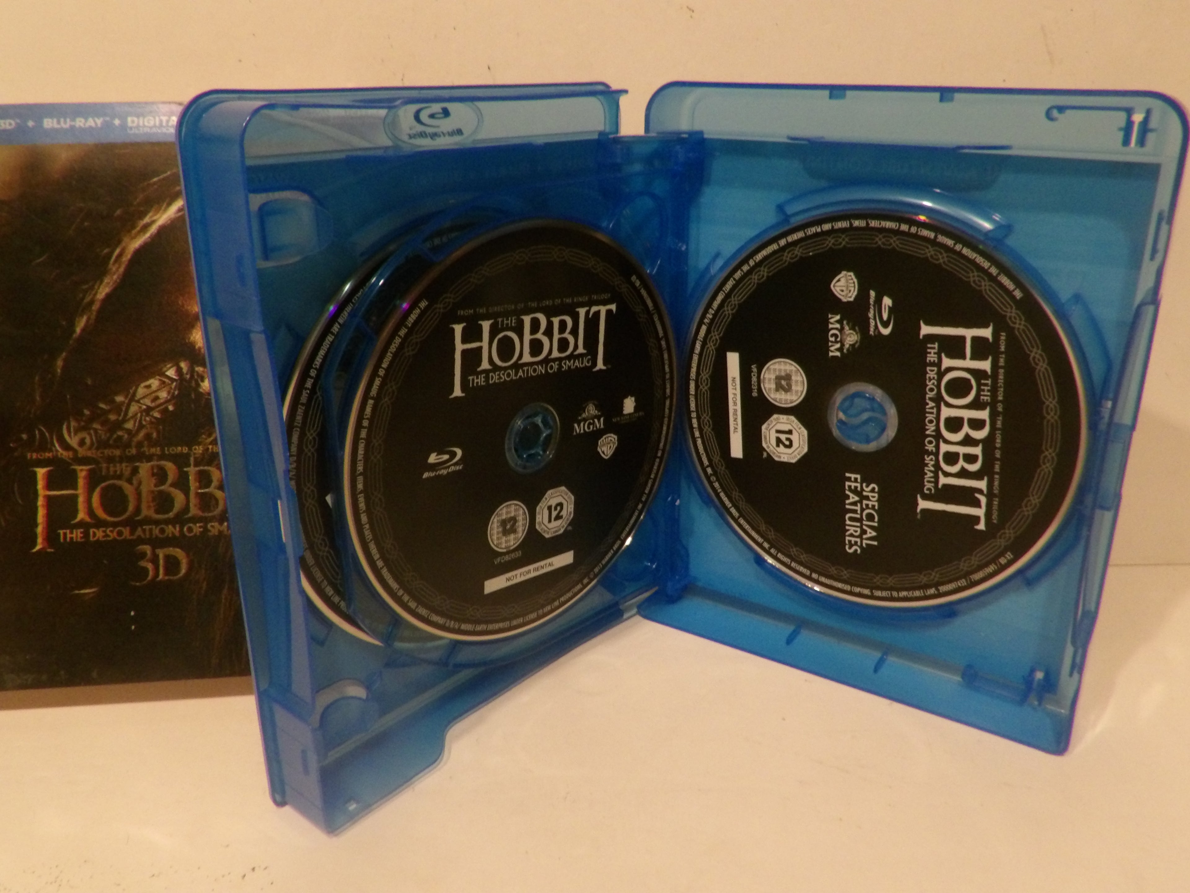 A blue plastic Blu-ray case open to show two discs for The Hobbit: The Desolation of Mordor 3D Blu-ray, with the case standing upright on a white surface and a box partially visible behind it.